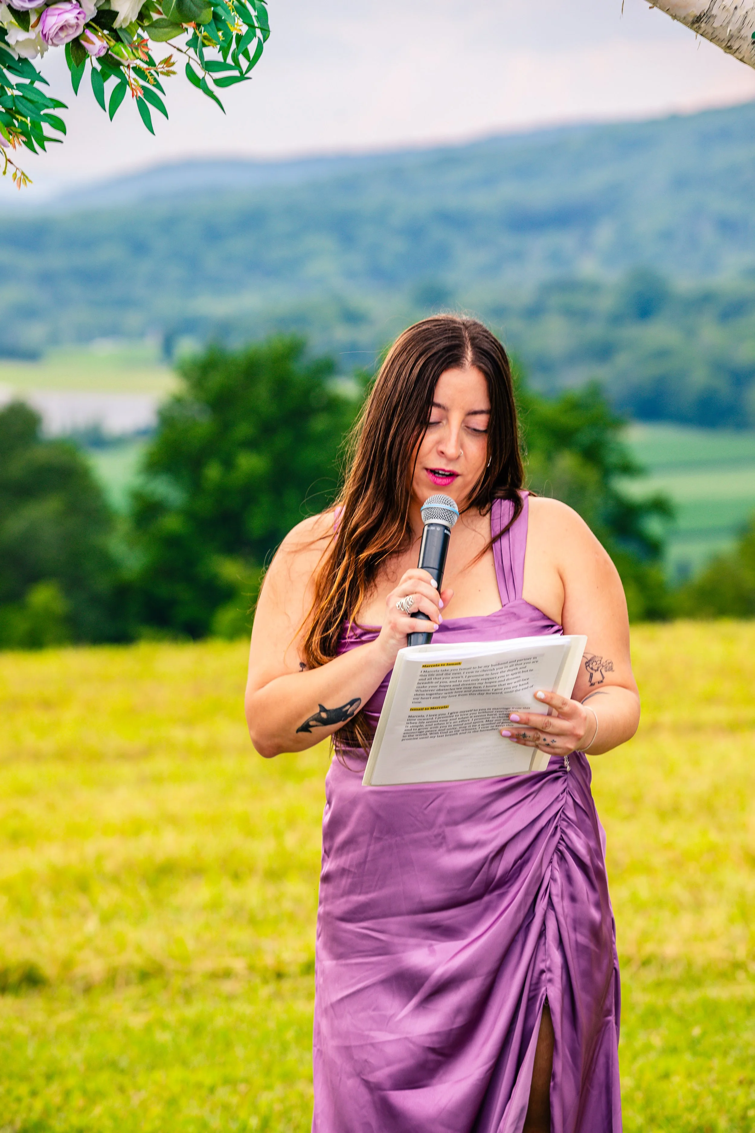 A woman in a lavender dress softly reading from a paper while holding a microphone in an outdoor setting with green hills and trees in the background.