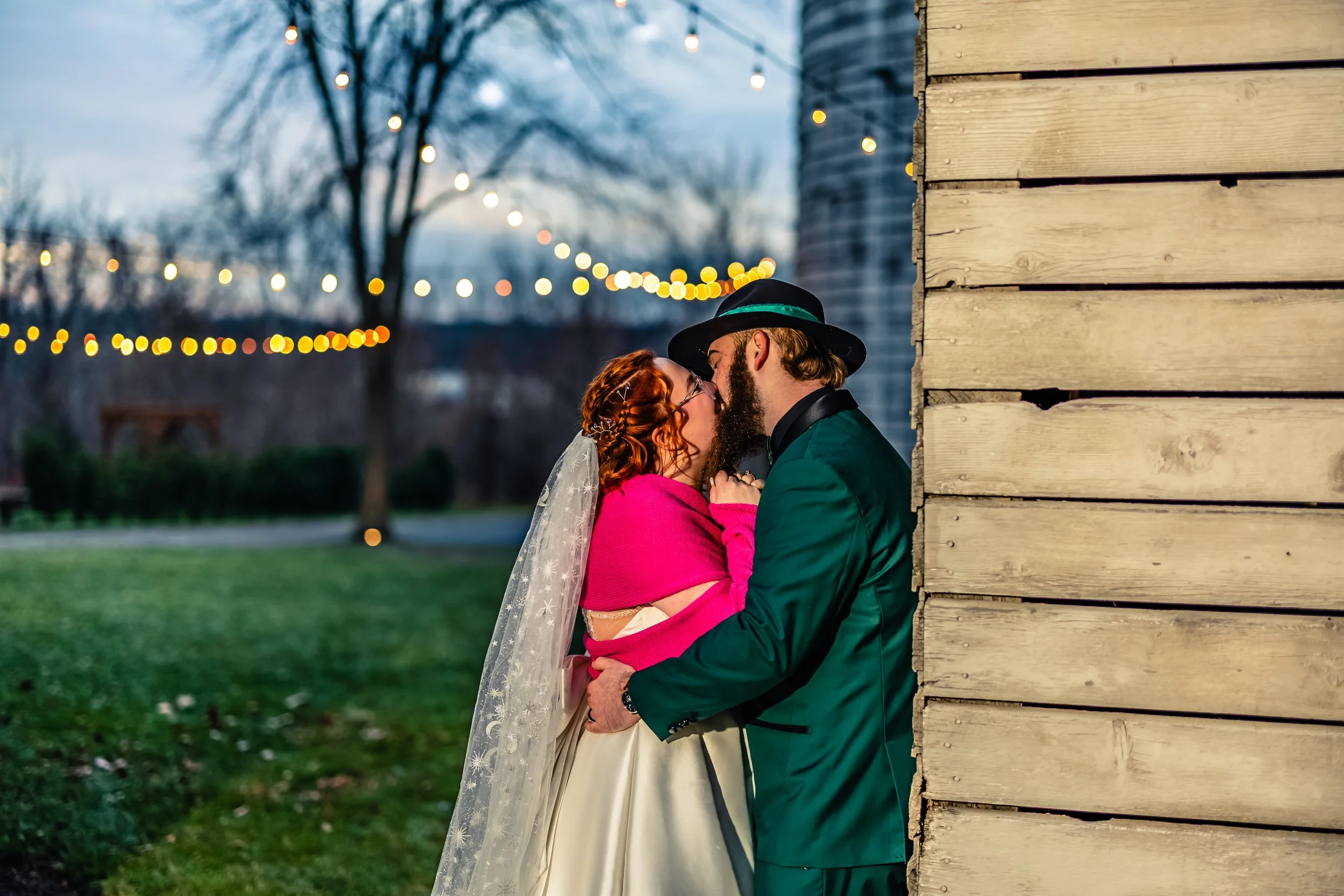 A couple dressed in wedding attire sharing a kiss outdoors during evening with string lights in the background.