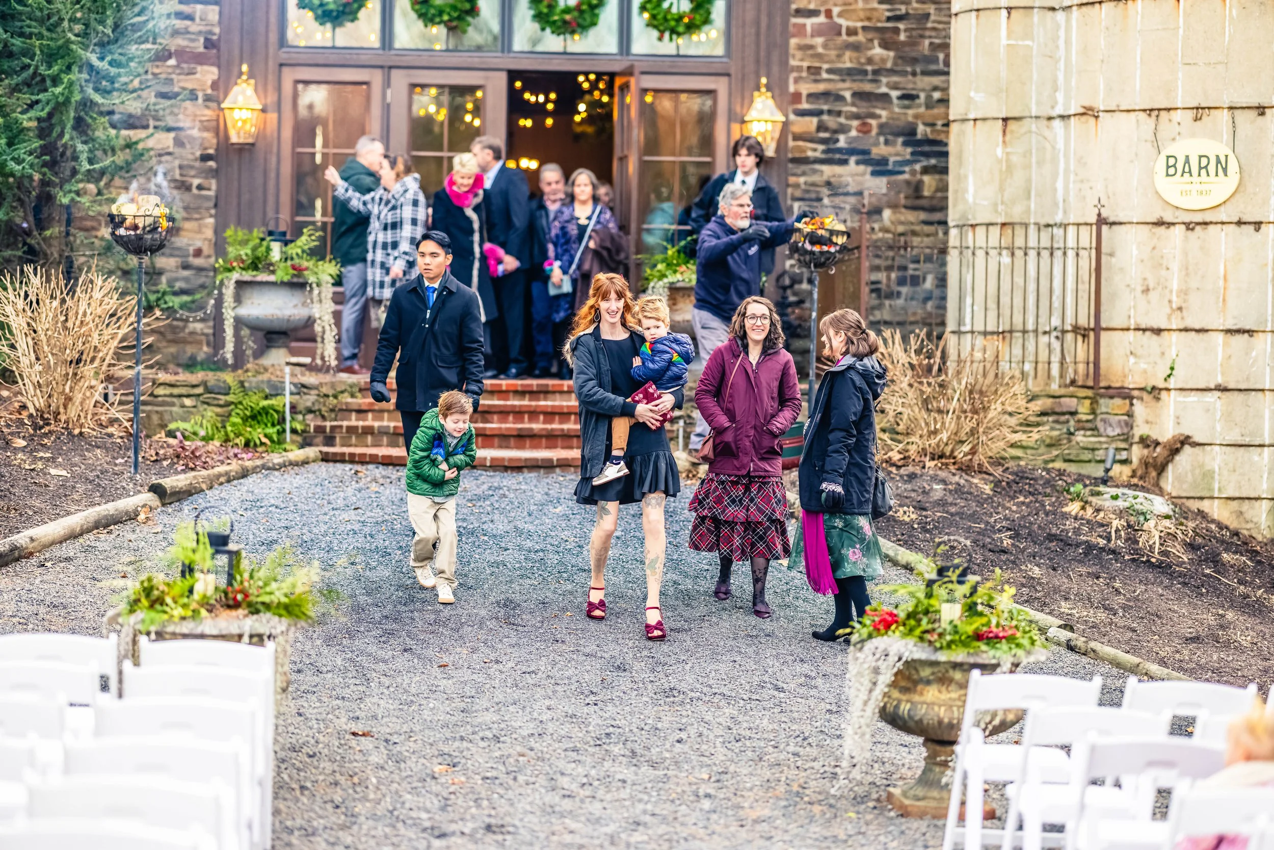 People gathered outside a stone building decorated for Christmas, some walking down a gravel path and others standing on steps, dressed in warm clothing.