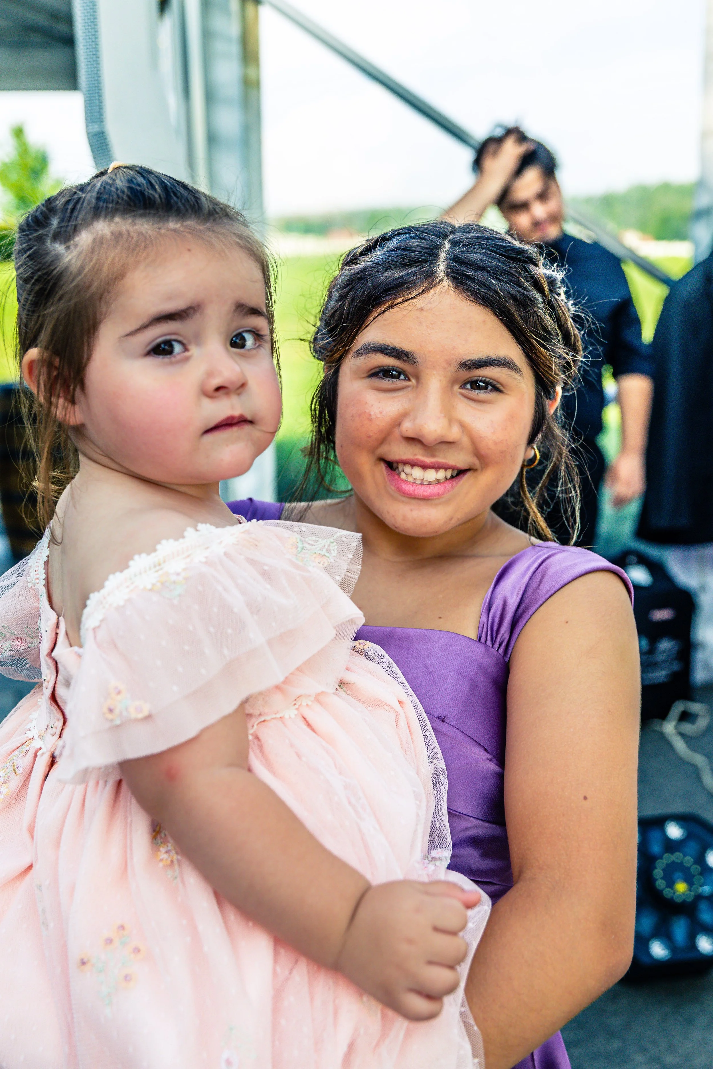 A young woman in a purple dress holding a small girl in a pink dress outdoors with a green field and two people in the background.