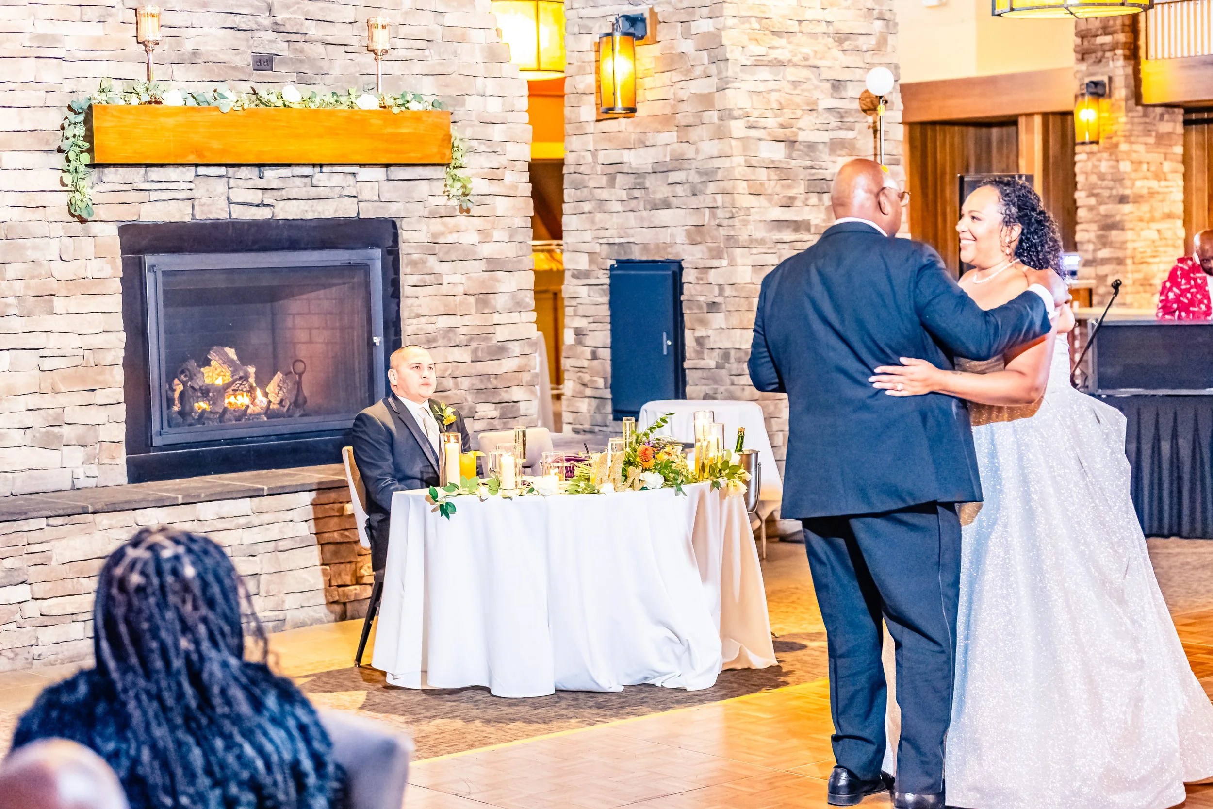 A couple dancing at a wedding reception, with a fireplace and guest table with candles in the background.
