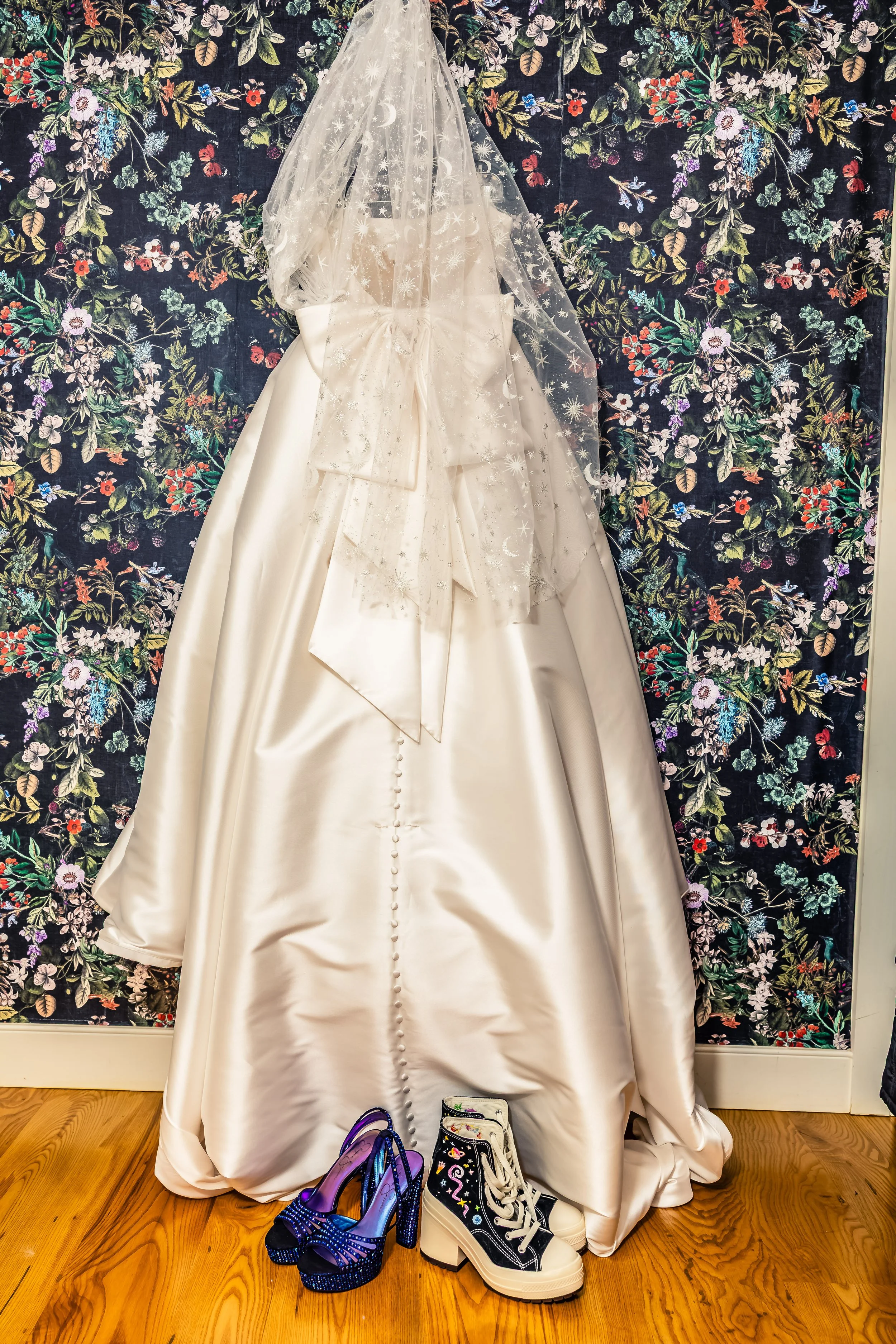 A wedding dress displayed on a mannequin with a veil, next to a pair of purple high-heeled shoes and a pair of black and white embroidered combat boots, set against a floral wallpaper background.