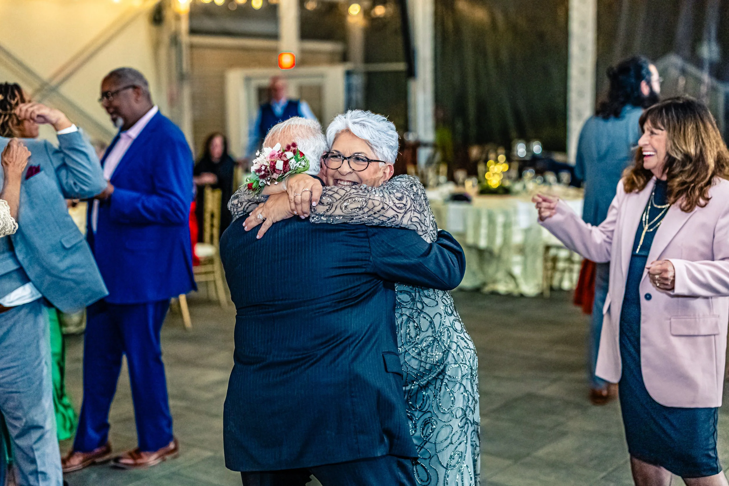 A joyful elderly woman with gray hair and glasses hugging an elderly man at a celebration or wedding, surrounded by other smiling guests dancing and enjoying the event indoors.