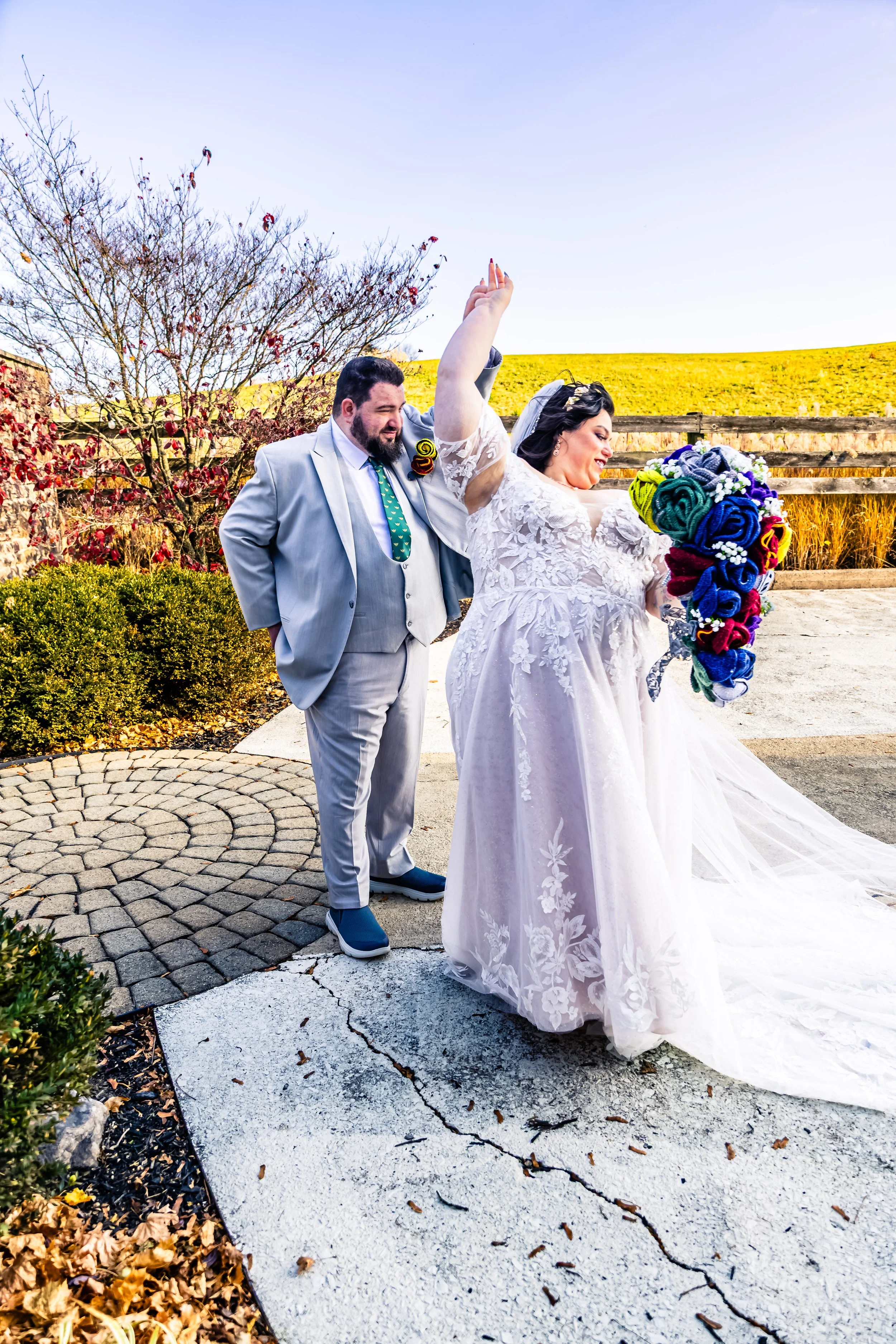 A bride in a white wedding dress holding a colorful bouquet, standing outside with a groom in a light gray suit looking on, on a bright day with autumn foliage in the background.