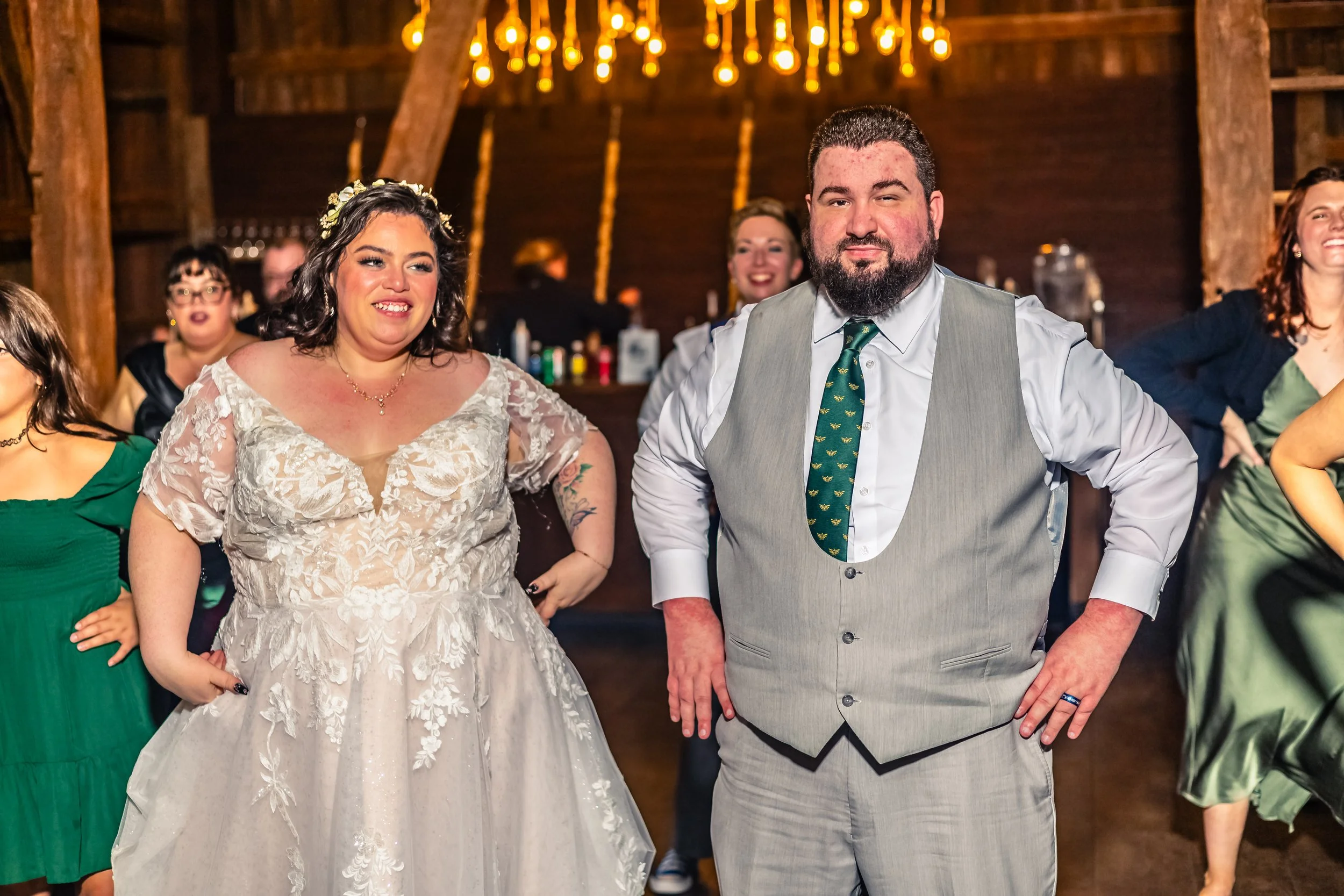 A group of people at a wedding reception, including a bride in a white lace wedding dress and a groom in a light grey suit with a green tie, standing indoors with wooden walls and warm lighting.