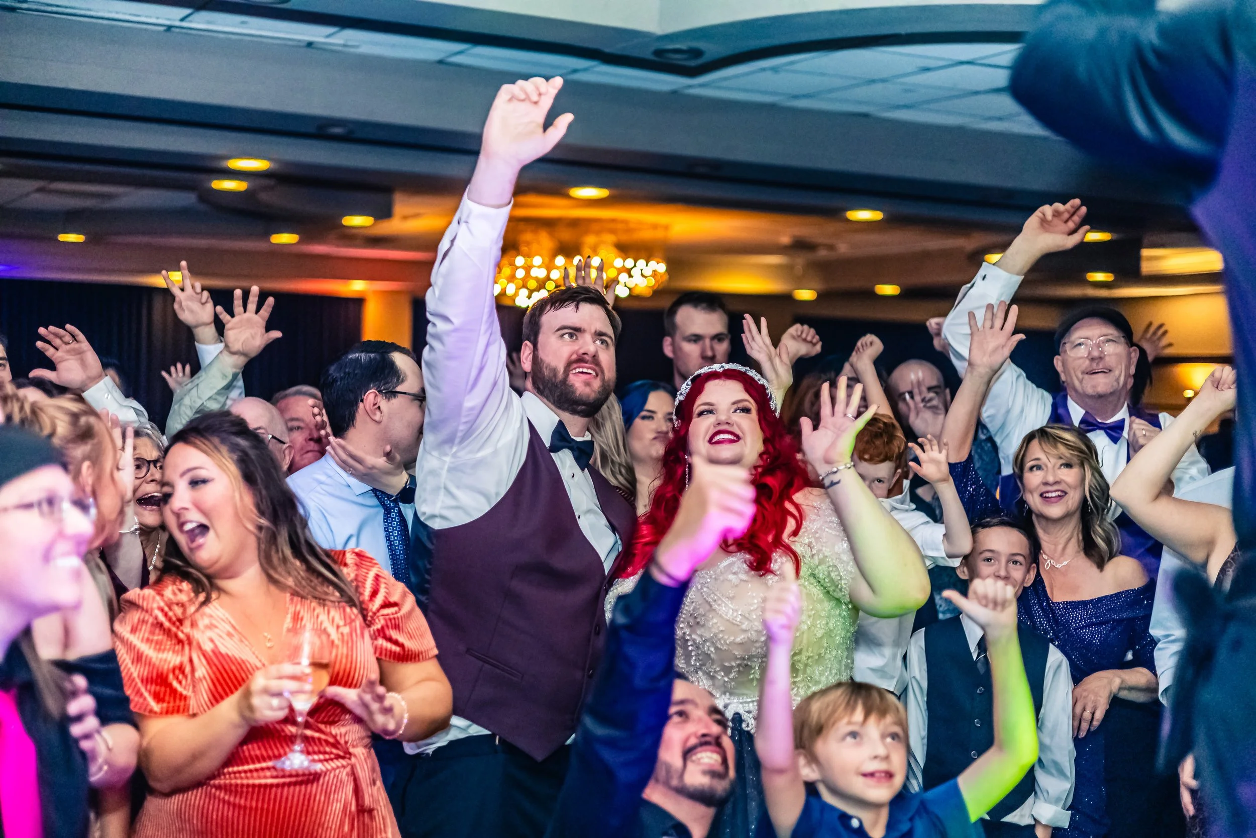 Crowd of people celebrating at a wedding reception with their hands raised, smiling and cheering.