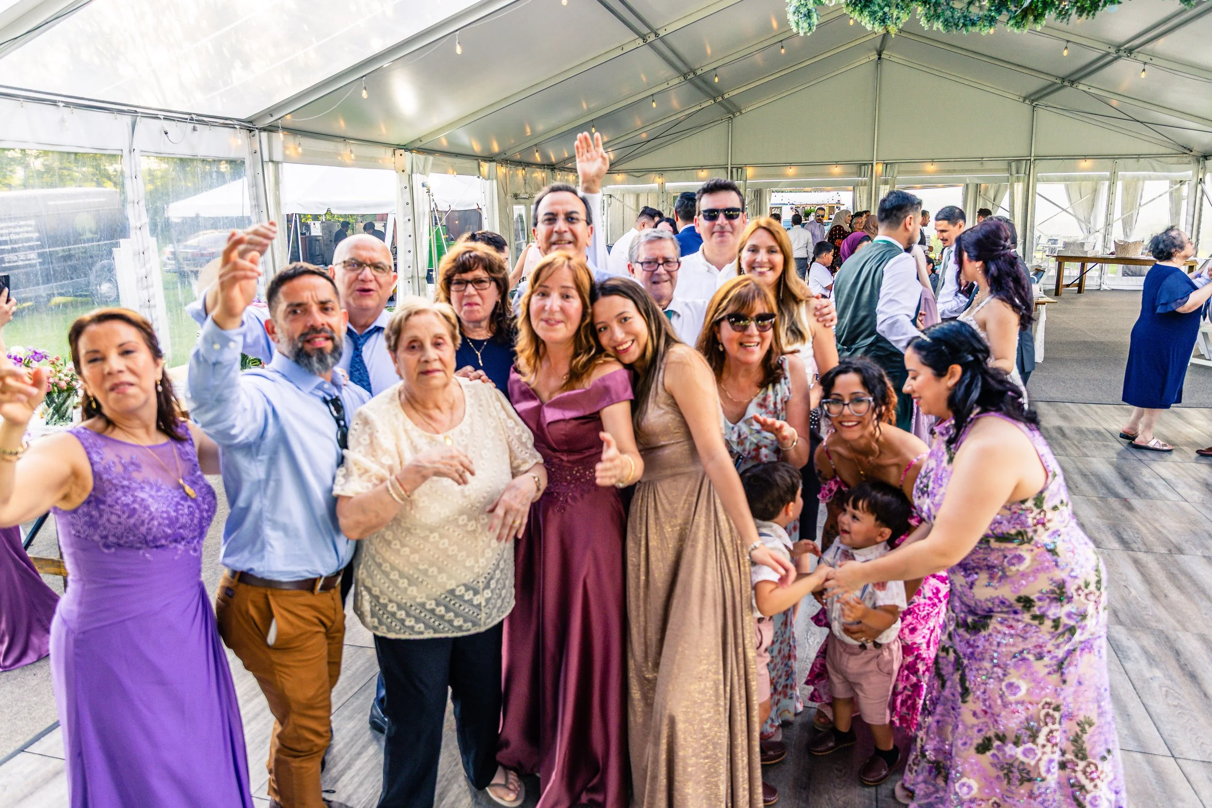 Group of people celebrating at a wedding under a large tent. They are dressed in formal and semi-formal attire, smiling, and posing for a photo.