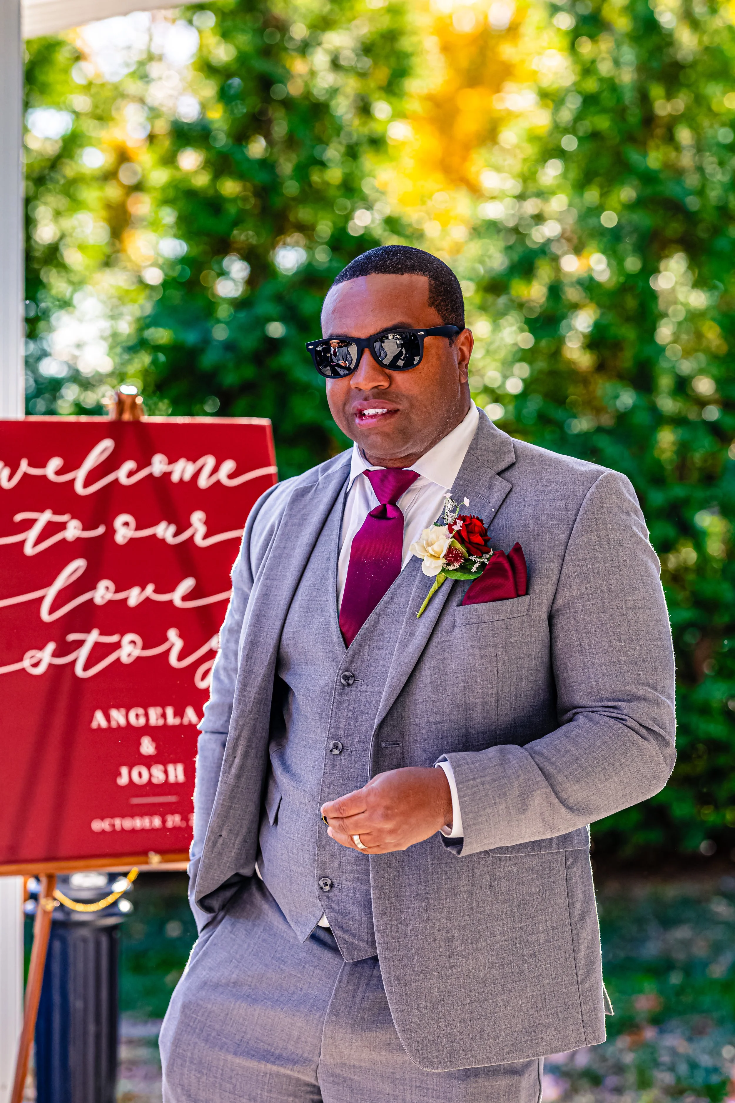A man in a gray suit with a white shirt and purple tie, wearing sunglasses and a boutonniere, standing outdoors at a wedding reception with a red welcome sign in the background.