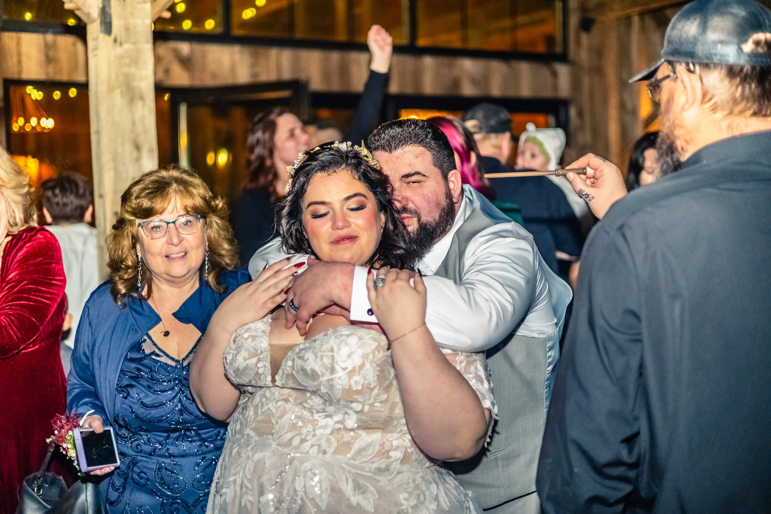 A group of people celebrating at a wedding reception, with a bride and groom hugging. The bride has dark hair and is wearing a lace wedding dress, while the groom has dark hair, a beard, and is wearing a light gray vest over a white shirt. A woman in