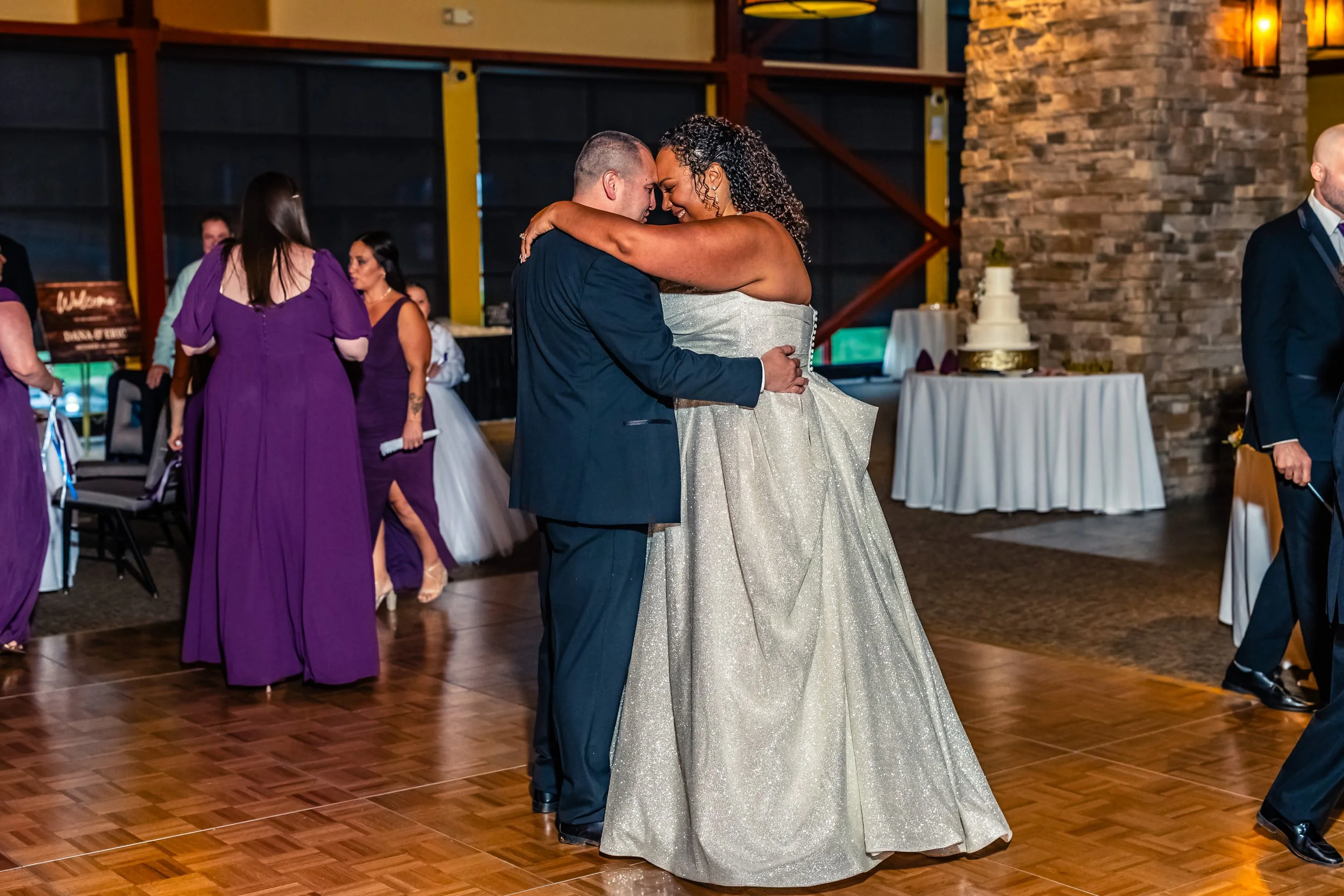 A bride and groom dancing closely at their wedding reception, with wedding guests in the background and a wedding cake on a table to the side.