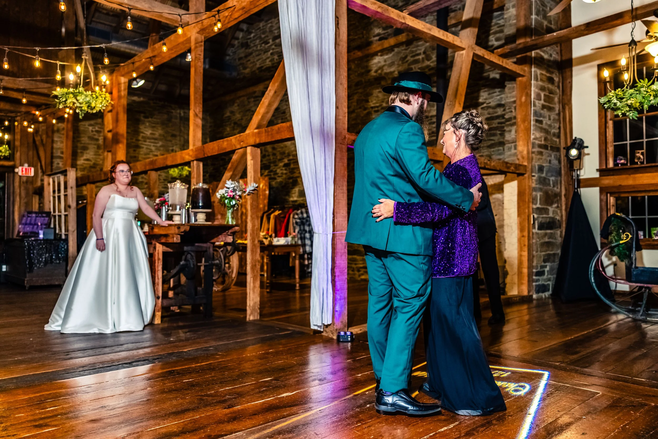 Couple dancing at a wedding reception in a rustic venue with wooden beams and stone walls, while a woman in a white dress watches.