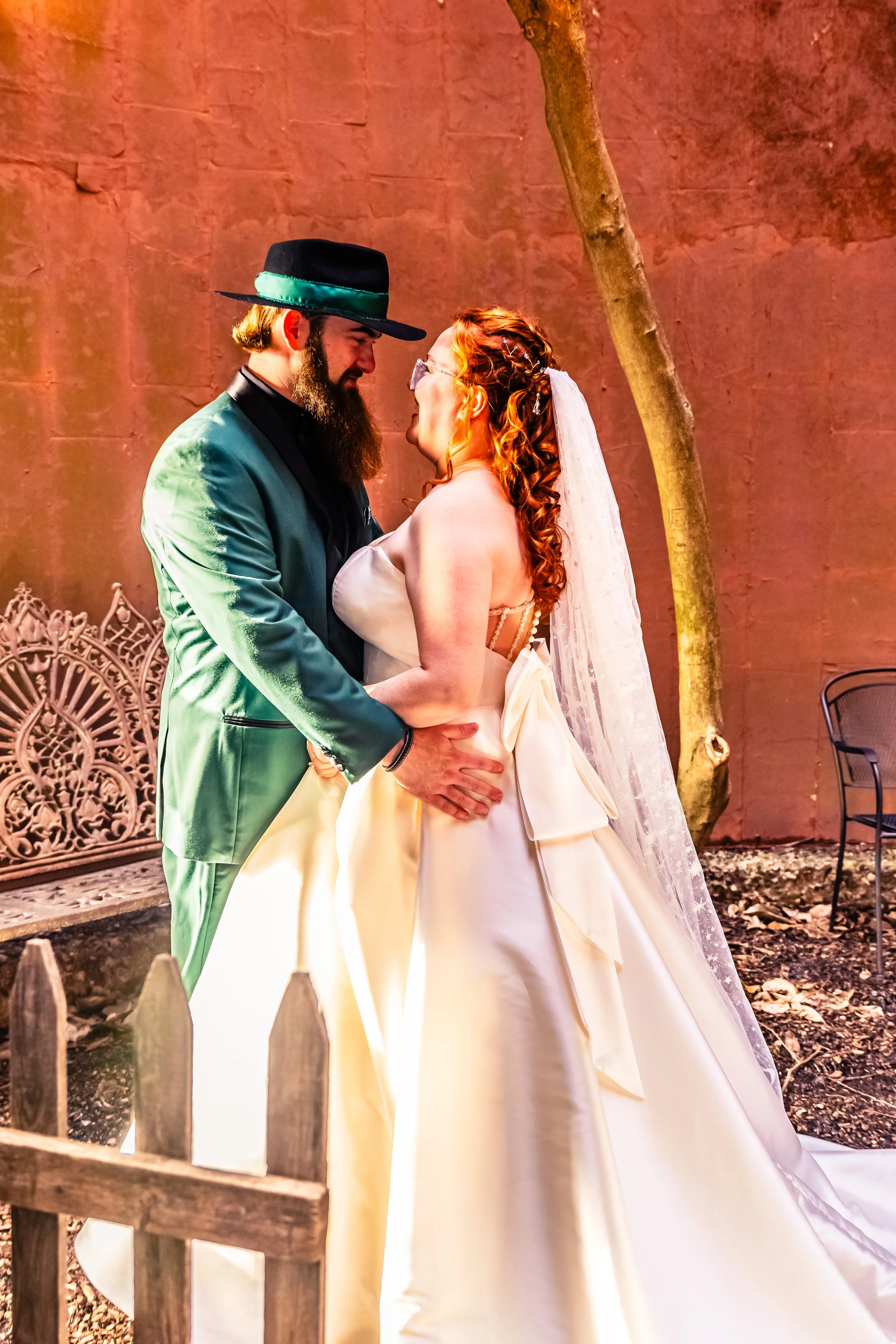 A couple sharing a kiss at a wedding reception, the groom in a black hat and suit, the bride in a white wedding dress with a long veil, outdoors with a tree and garden furniture in the background.