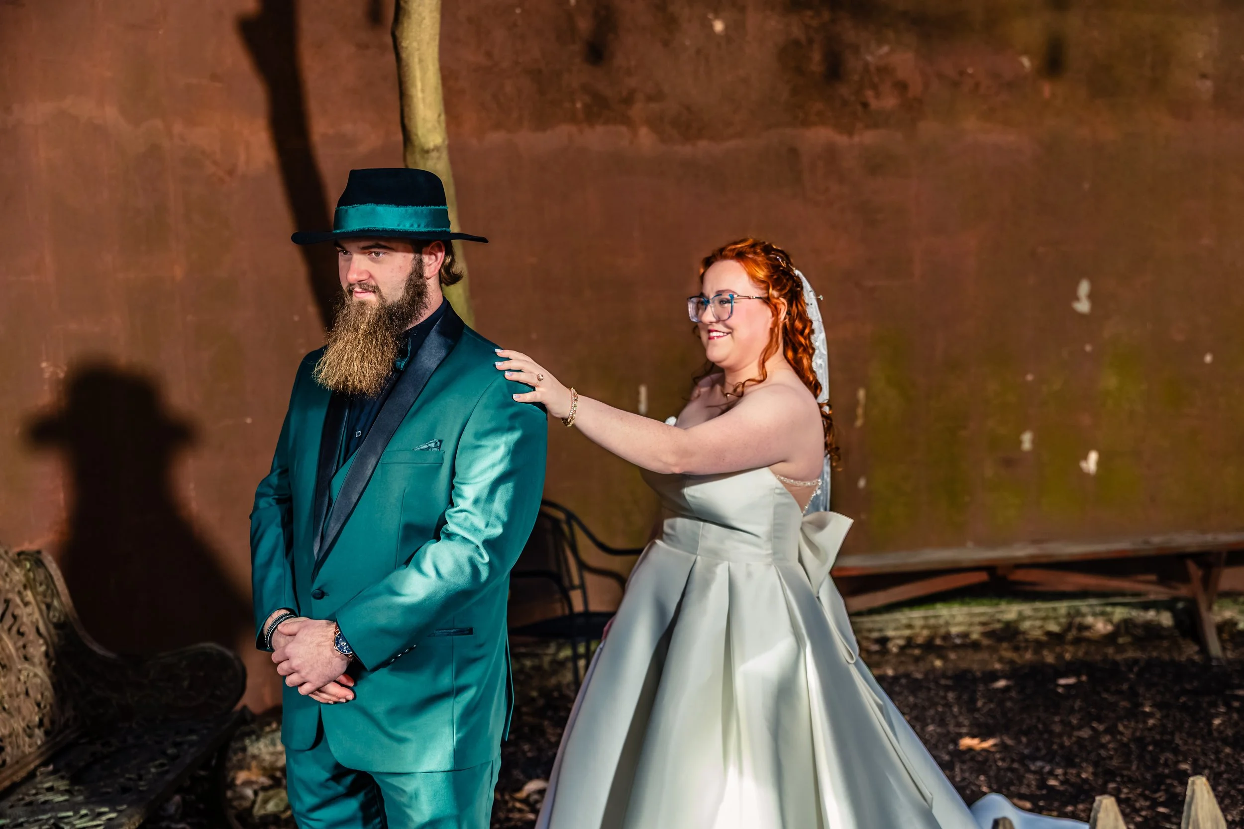 A bride and groom at their wedding, with the bride playfully pushing the groom's shoulder from behind as they smile. The groom wears a teal suit and a fedora hat, and the bride wears a strapless white wedding gown with a large bow at the back.