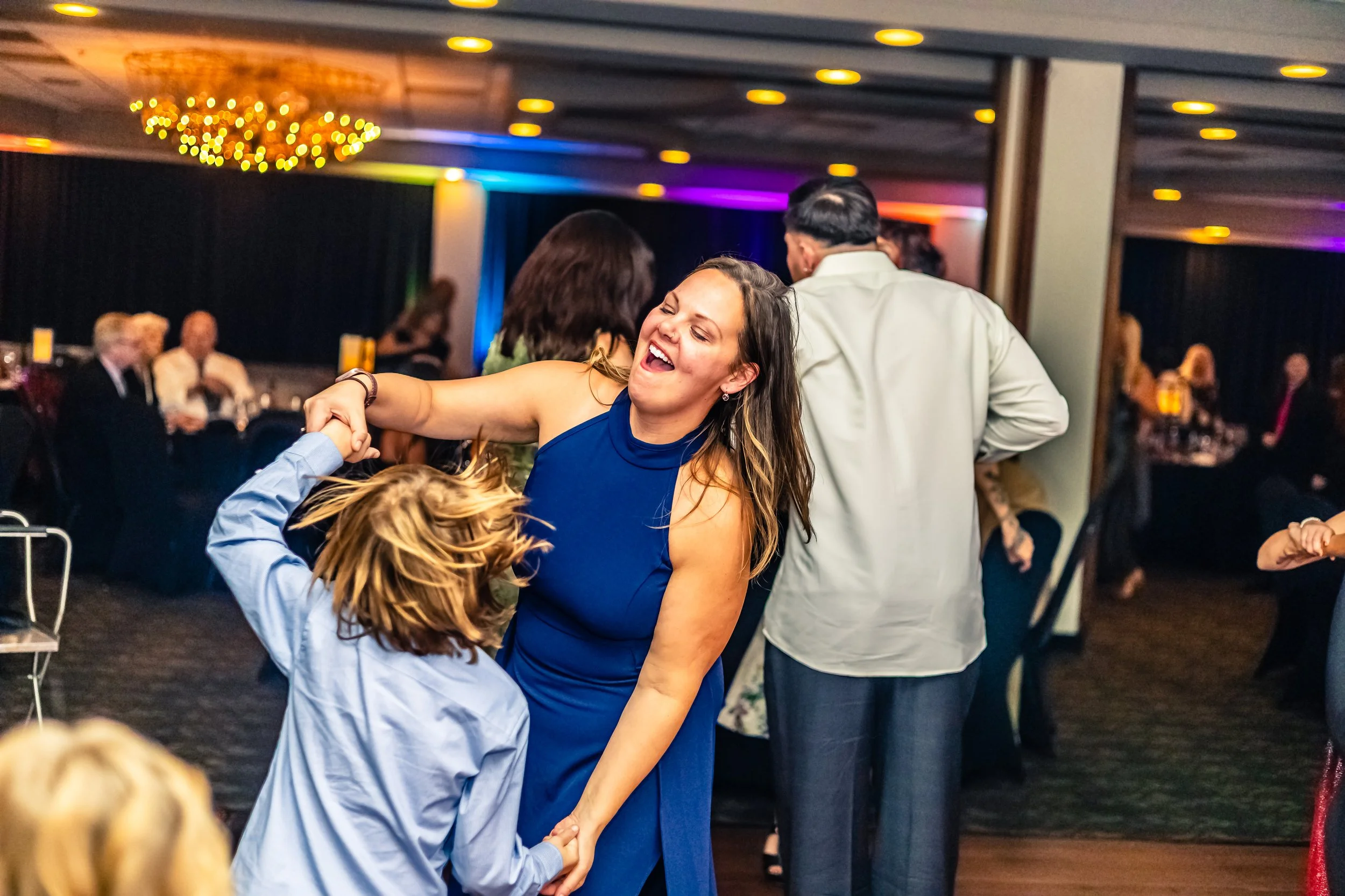 A woman and a young girl dance together at a party, holding hands and smiling. The woman is wearing a blue dress, and the girl is wearing a light blue shirt. People are visible in the background on a dance floor and seated at tables, with colorful li