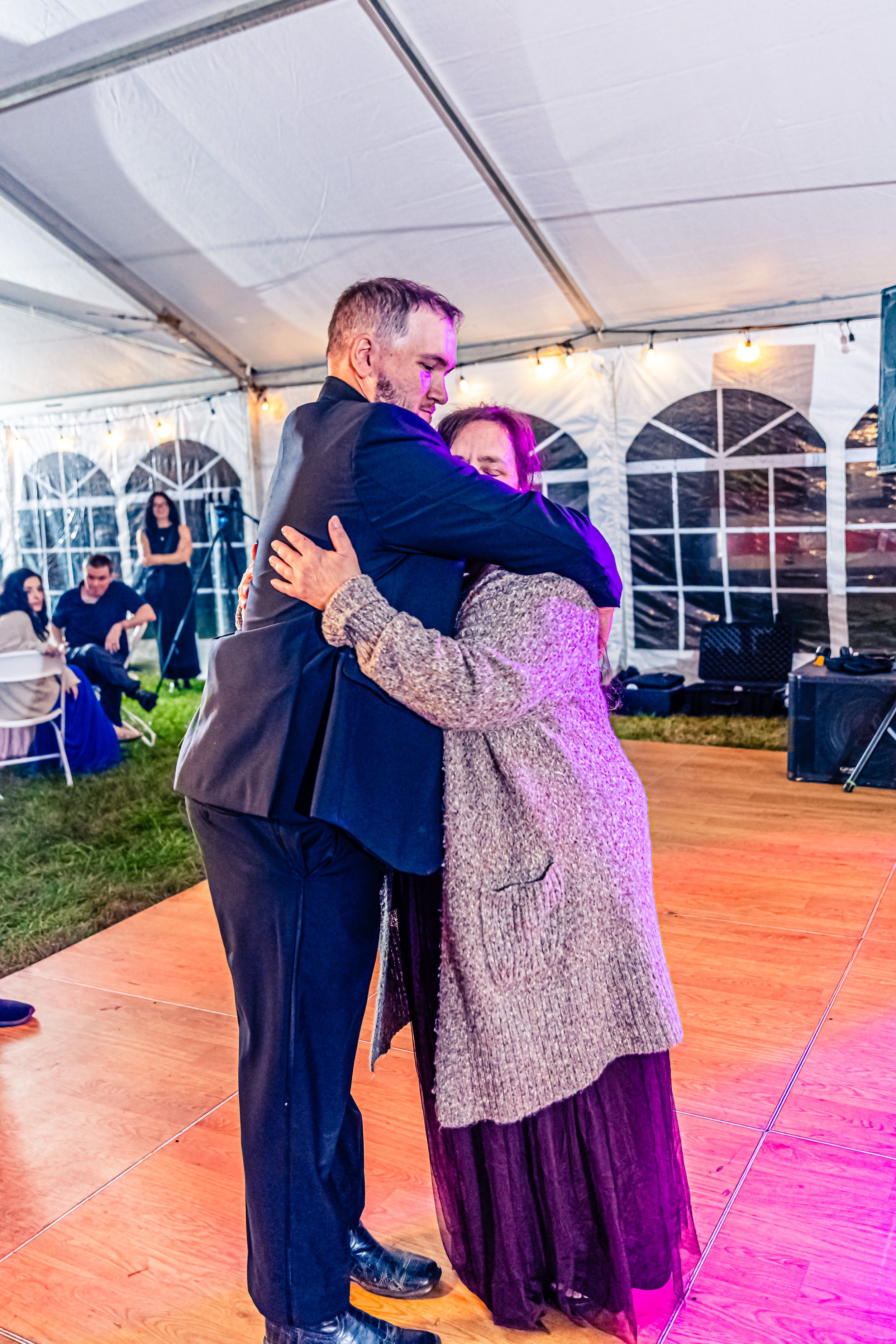 A man and woman are hugging on a dance floor at a celebration inside a decorated tent with string lights.