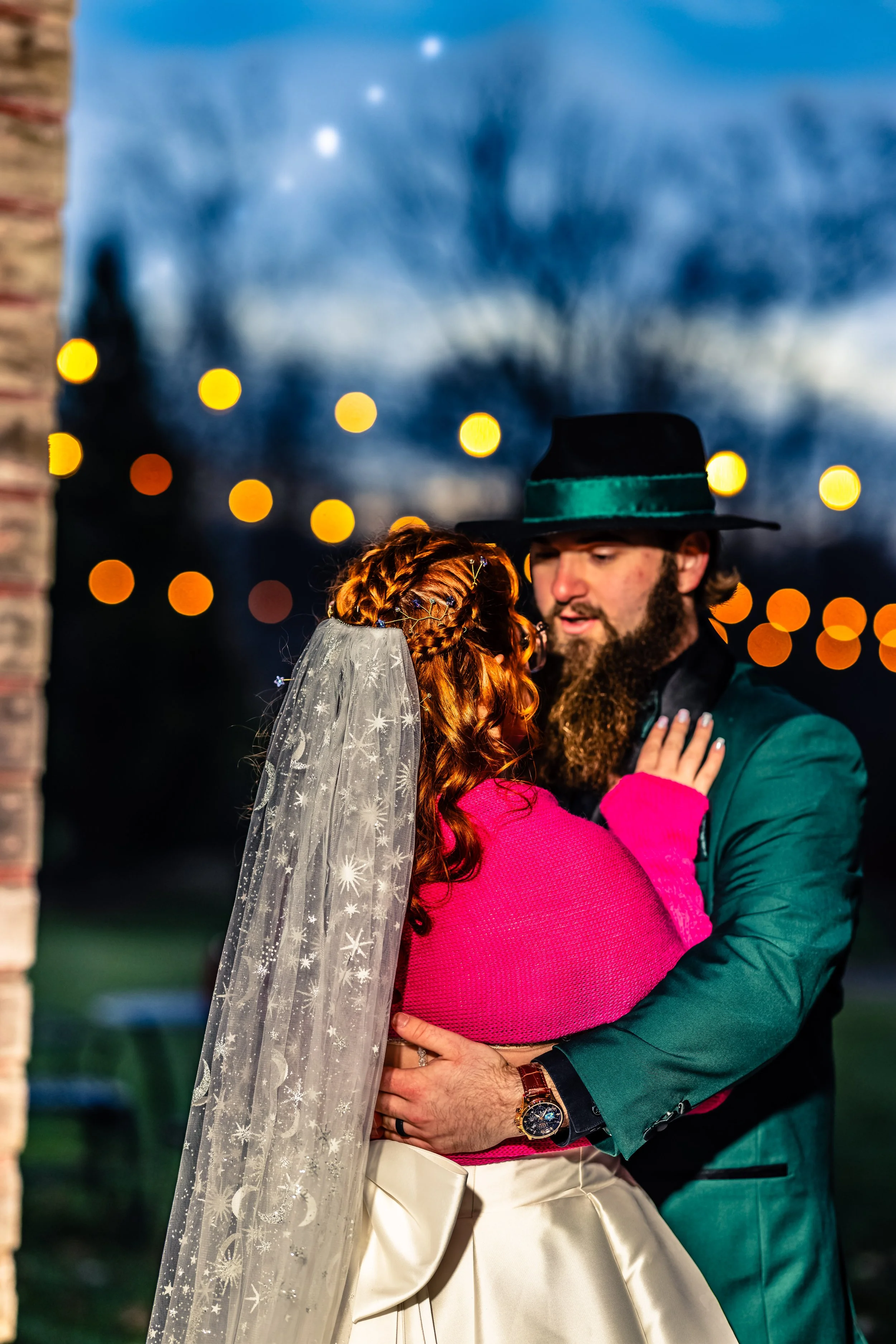 A couple dancing closely outdoors during evening, the woman with long red hair and a lace veil, wearing a pink top and beige skirt, and the man with a beard, wearing a teal jacket, black hat, and a watch.