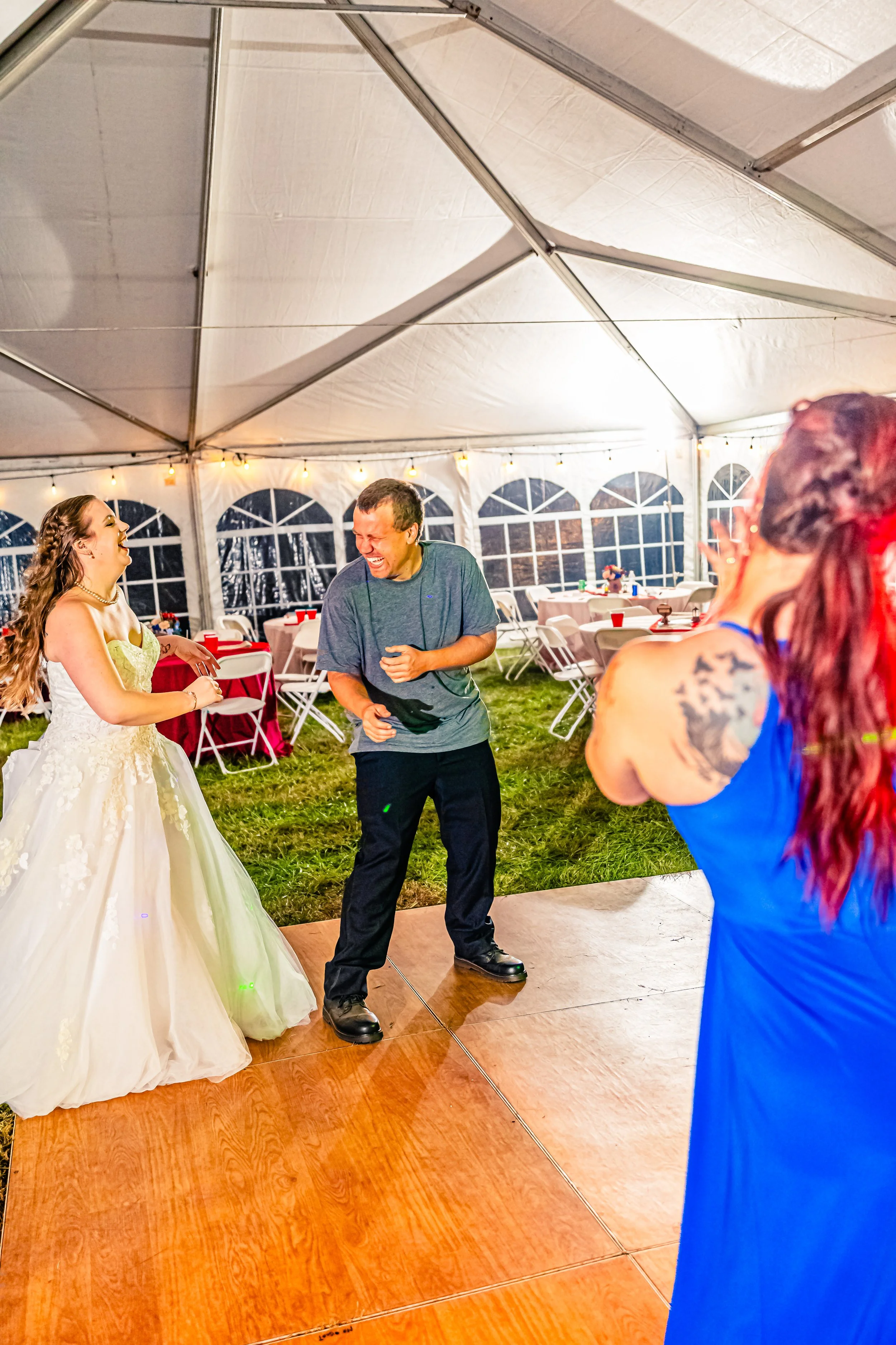 People dancing and laughing at a wedding reception inside a white tent with tables and string lights.