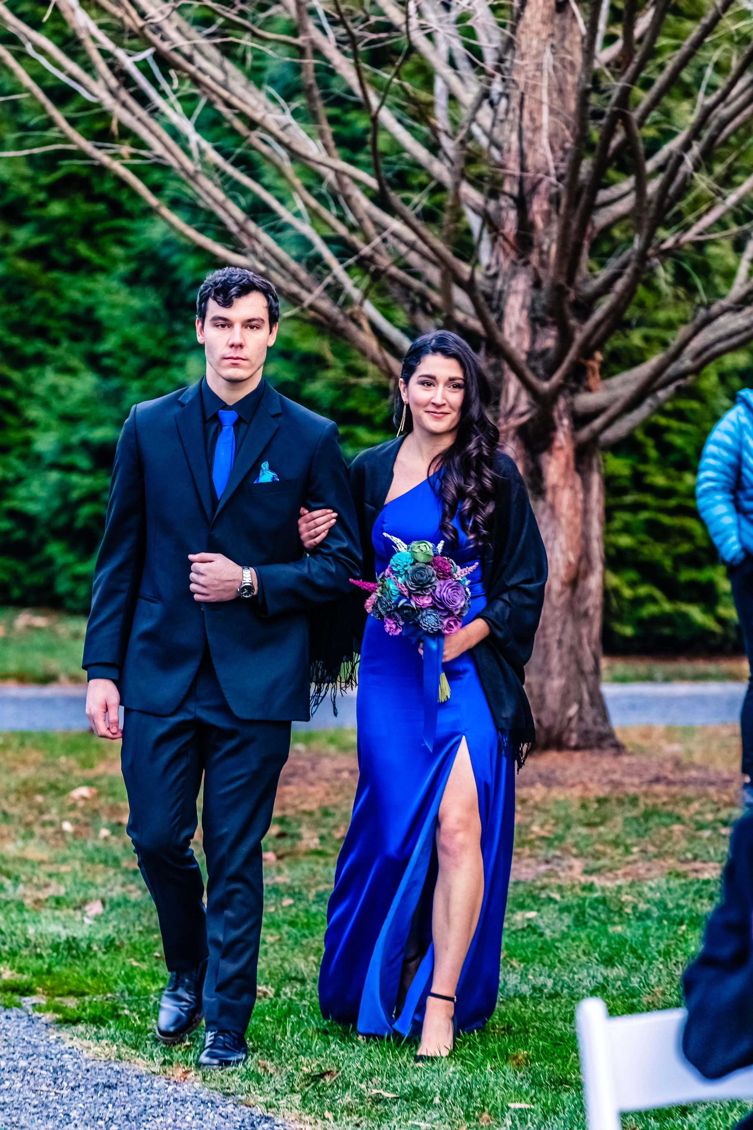 A couple walking outdoors, dressed in formal attire; the woman is holding a bouquet of flowers and wearing a blue gown with a high slit, and the man is in a black suit and blue tie, with a black shawl over his shoulders, walking arm-in-arm in front o