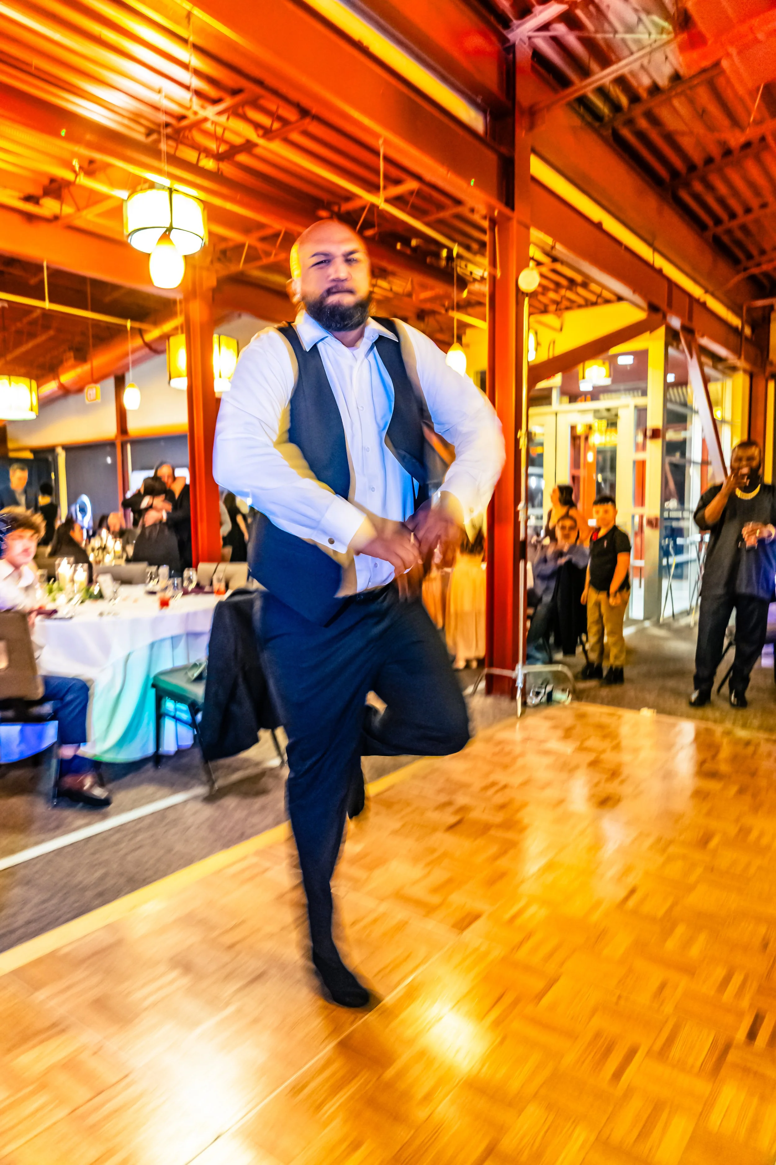 A man with a beard in a white shirt and dark vest dancing on a wooden floor at a celebration or event, with people seated and standing around in a warmly lit venue.