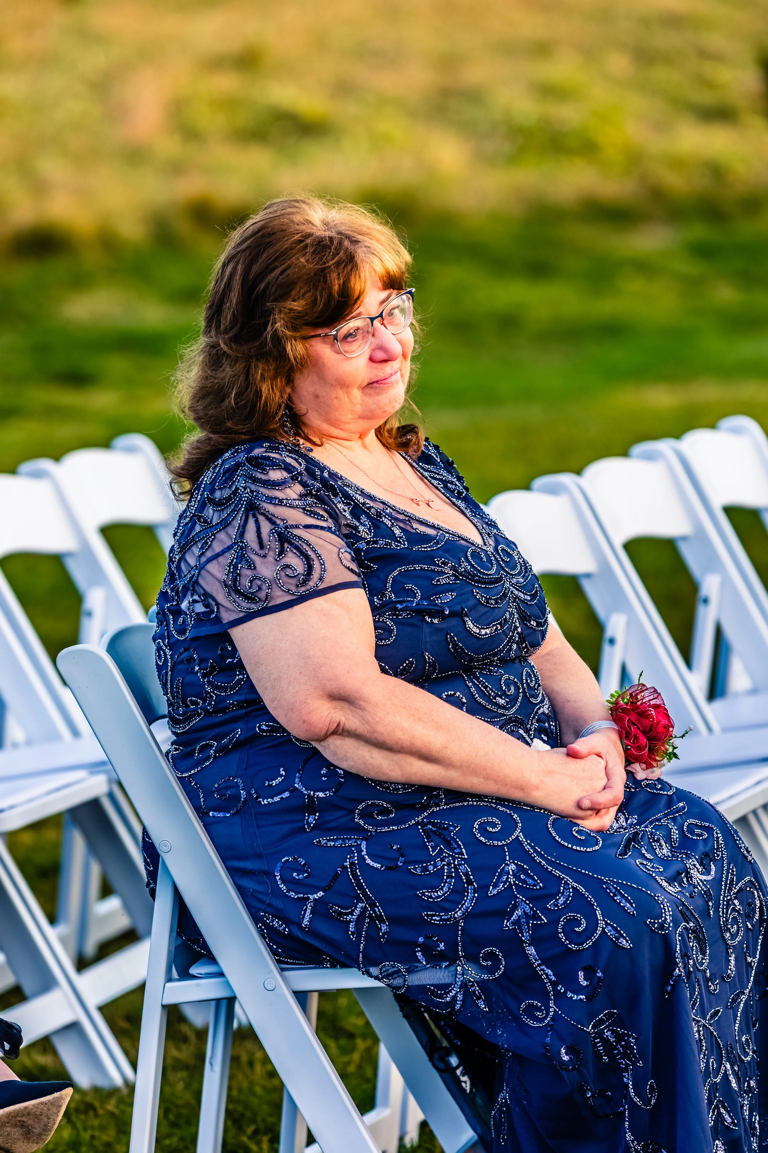 A woman in a navy blue, intricately beaded dress sitting outdoors on white folding chairs, holding a small bouquet of red roses, during what appears to be a formal event or wedding ceremony.