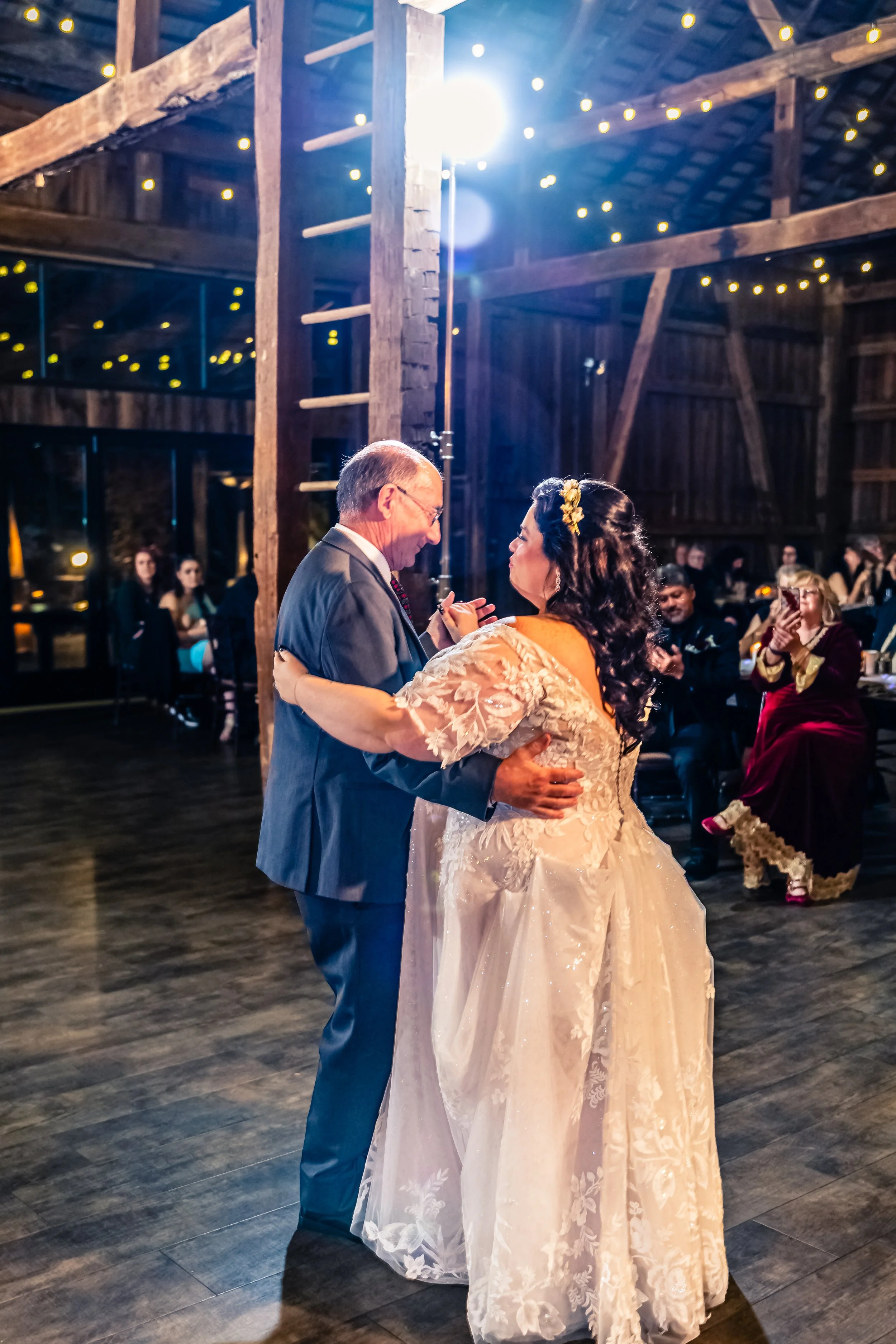 Bride and groom dancing at wedding reception in barn with guests seated at tables in background.