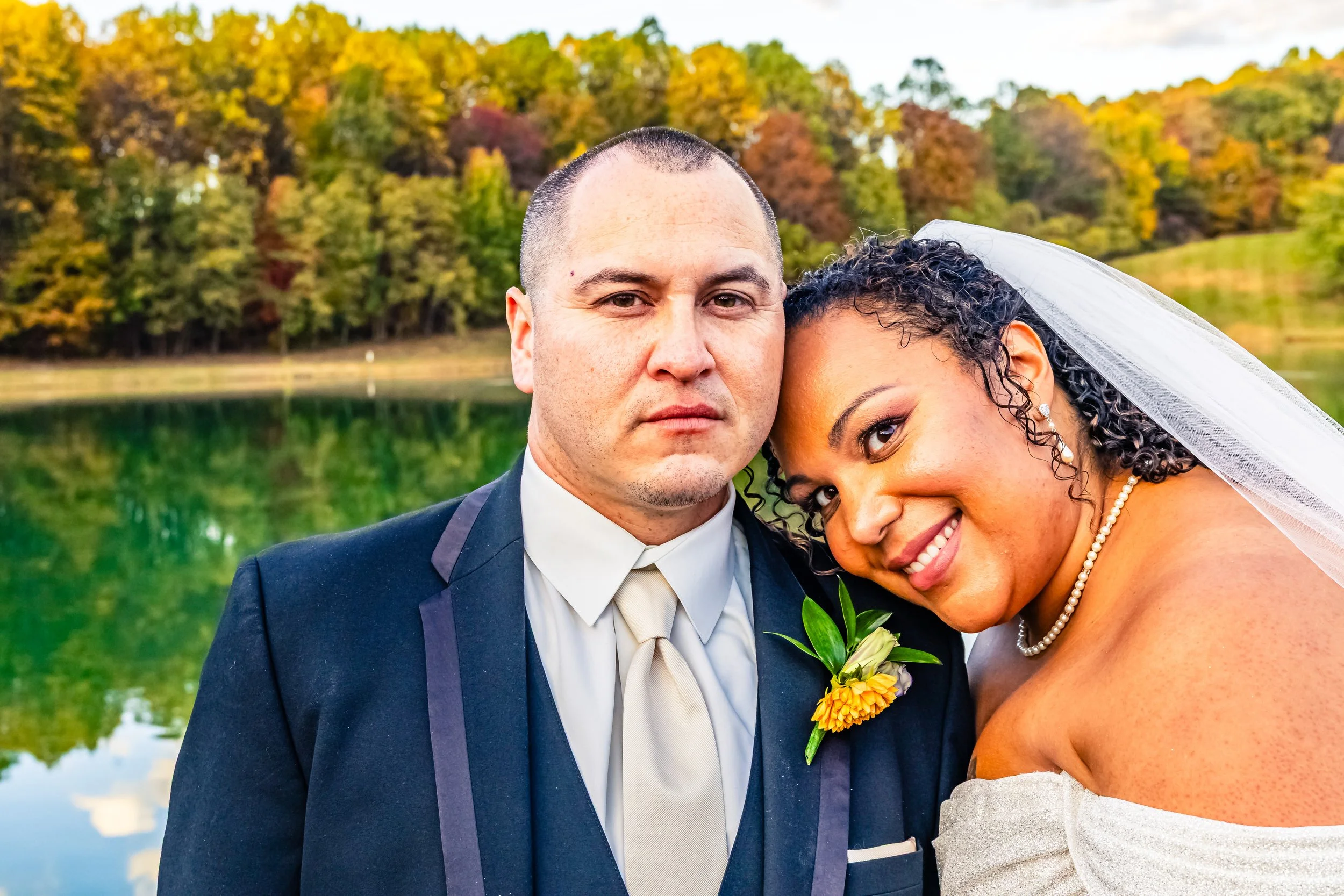 A newlywed couple takes a close-up selfie outdoors near a lake with autumn trees in the background. The groom is in a black suit with a cream tie and boutonniere, the bride is wearing a white wedding dress with a pearl necklace and veil, and they are