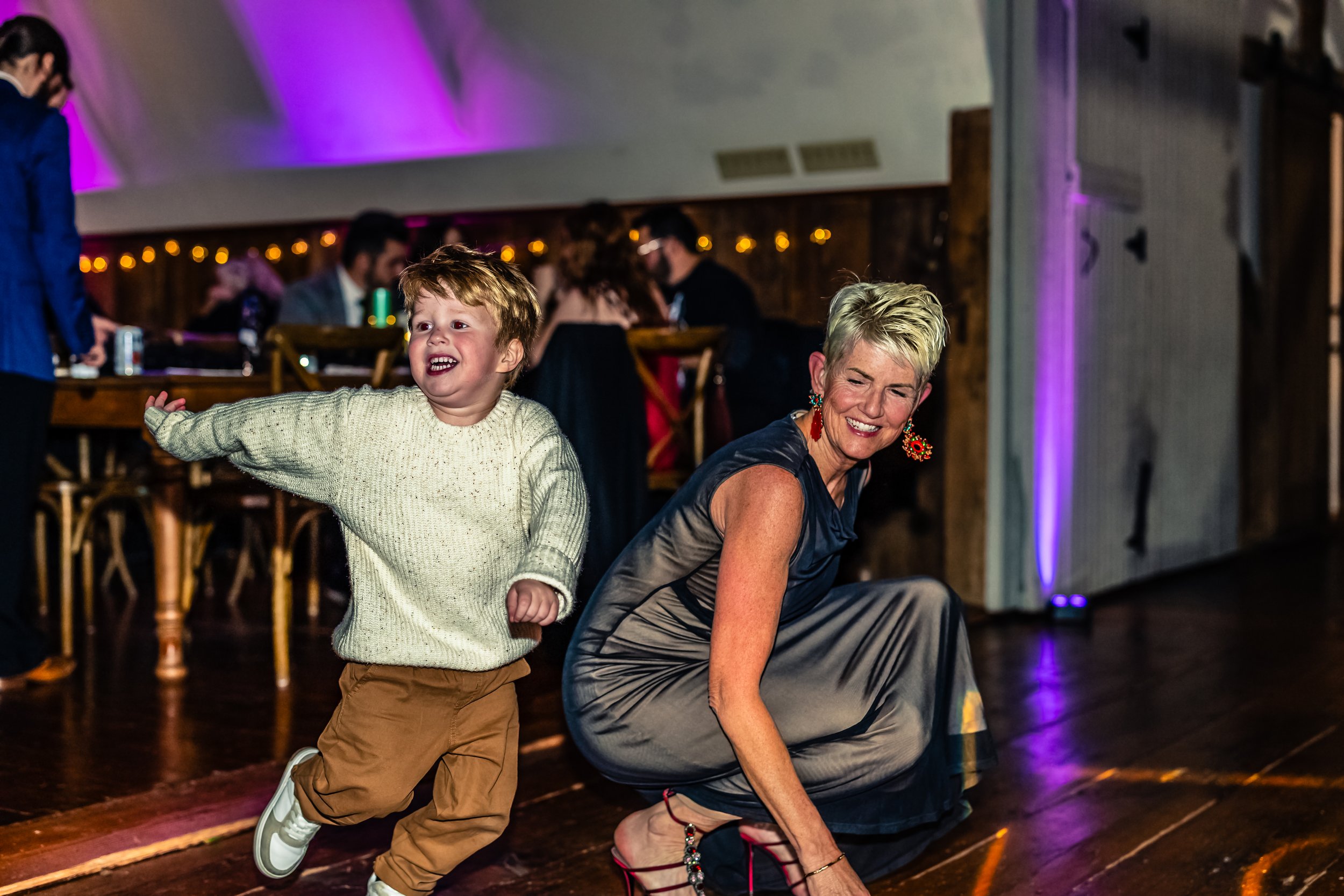 A young boy in a cream sweater and tan pants dancing happily with a smiling woman in a long gown at an indoor event with people sitting at tables in the background.