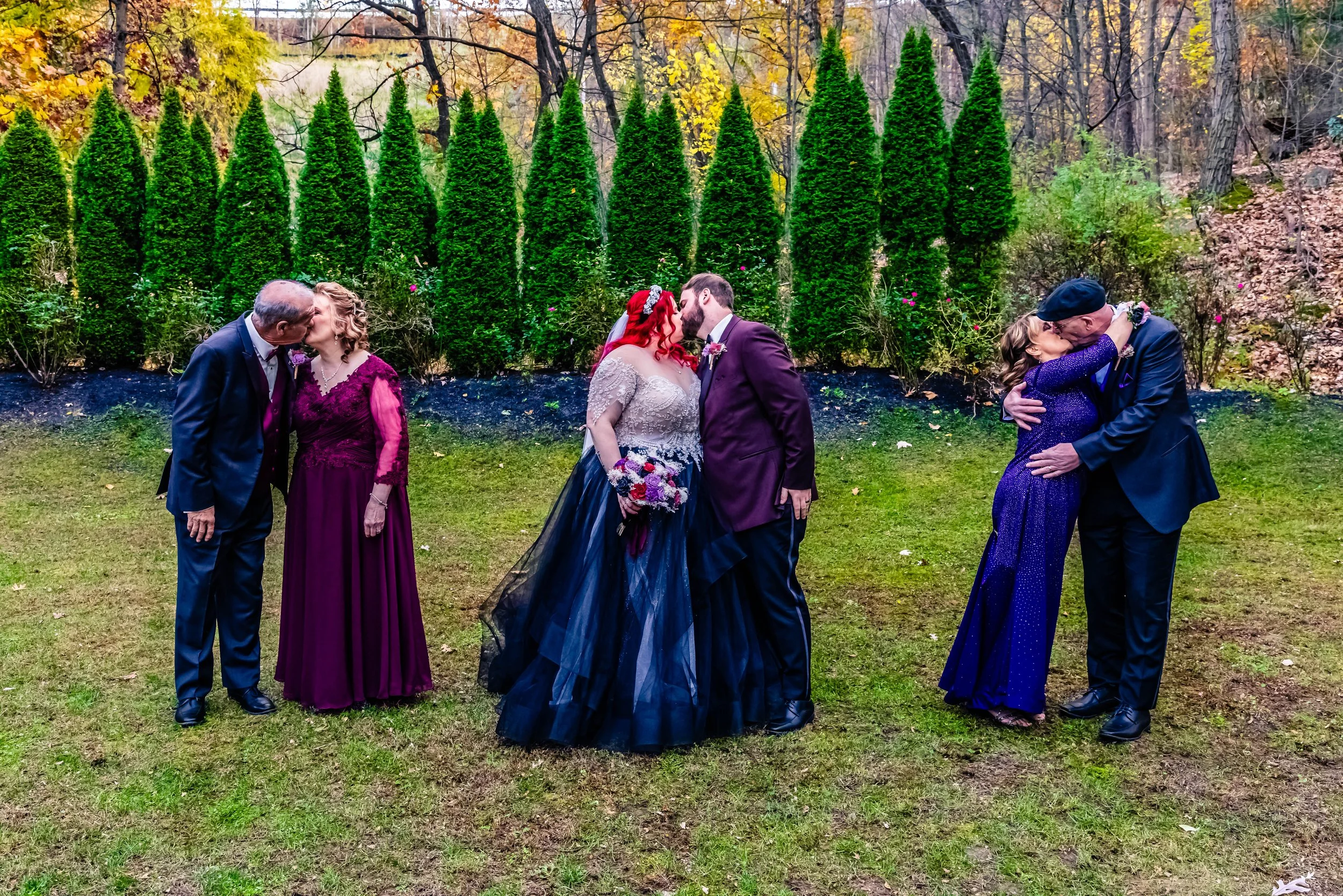 Group of couples kissing outdoors during a wedding, with trees and green grass in the background.