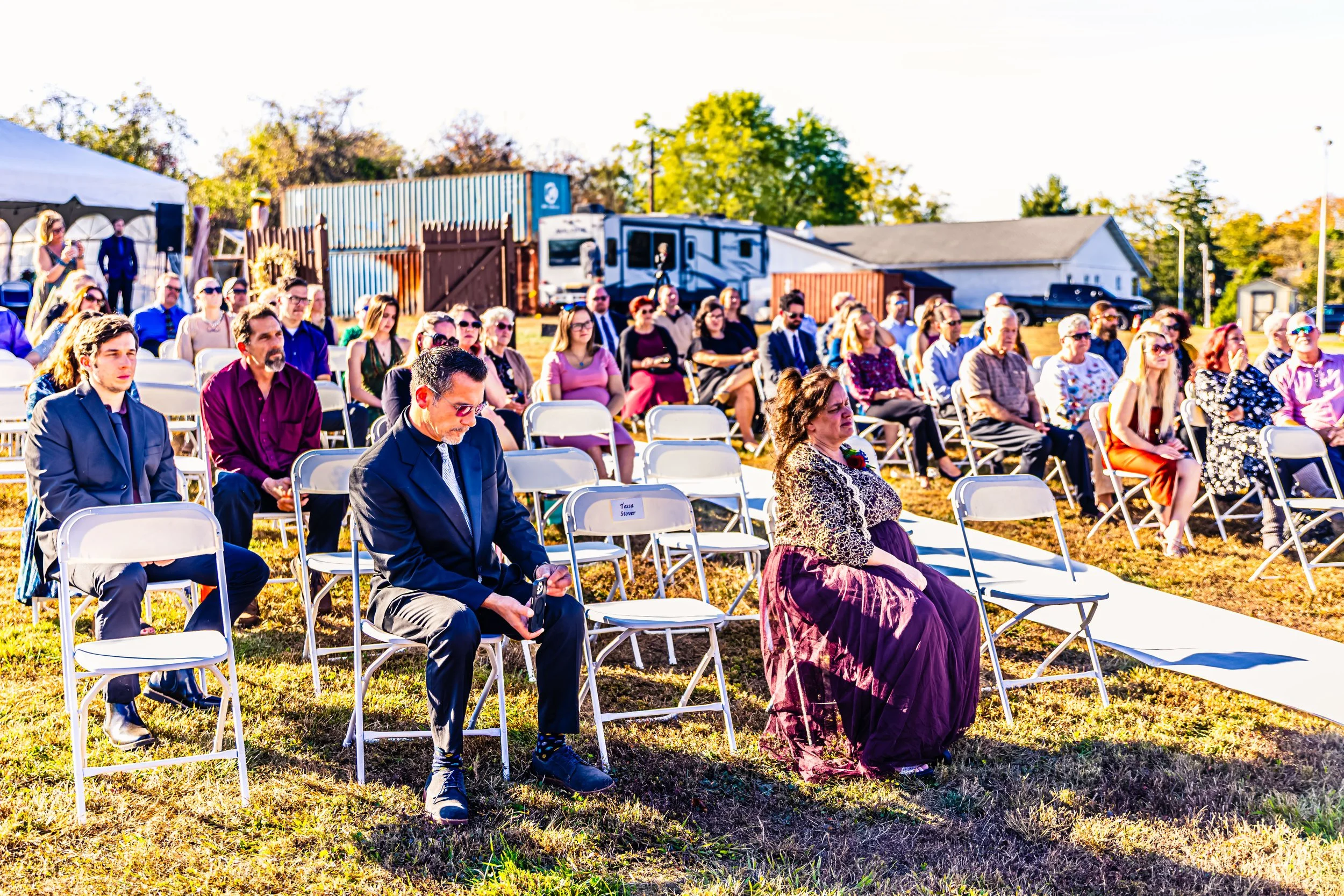 People seated outdoors on folding chairs at an event on a sunny day, with some standing in the background, on a grassy area with buildings and trees in the distance.