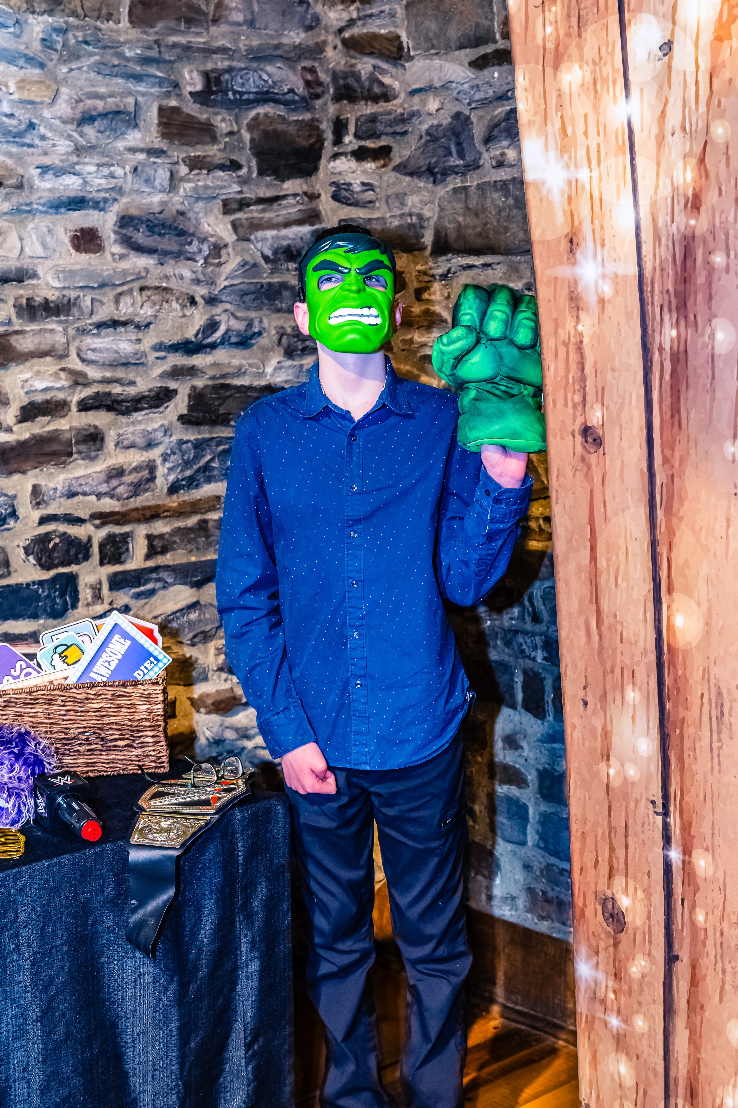 A person wearing a Hulk mask and a green Hulk glove, standing next to a curtain in a brick-walled room.