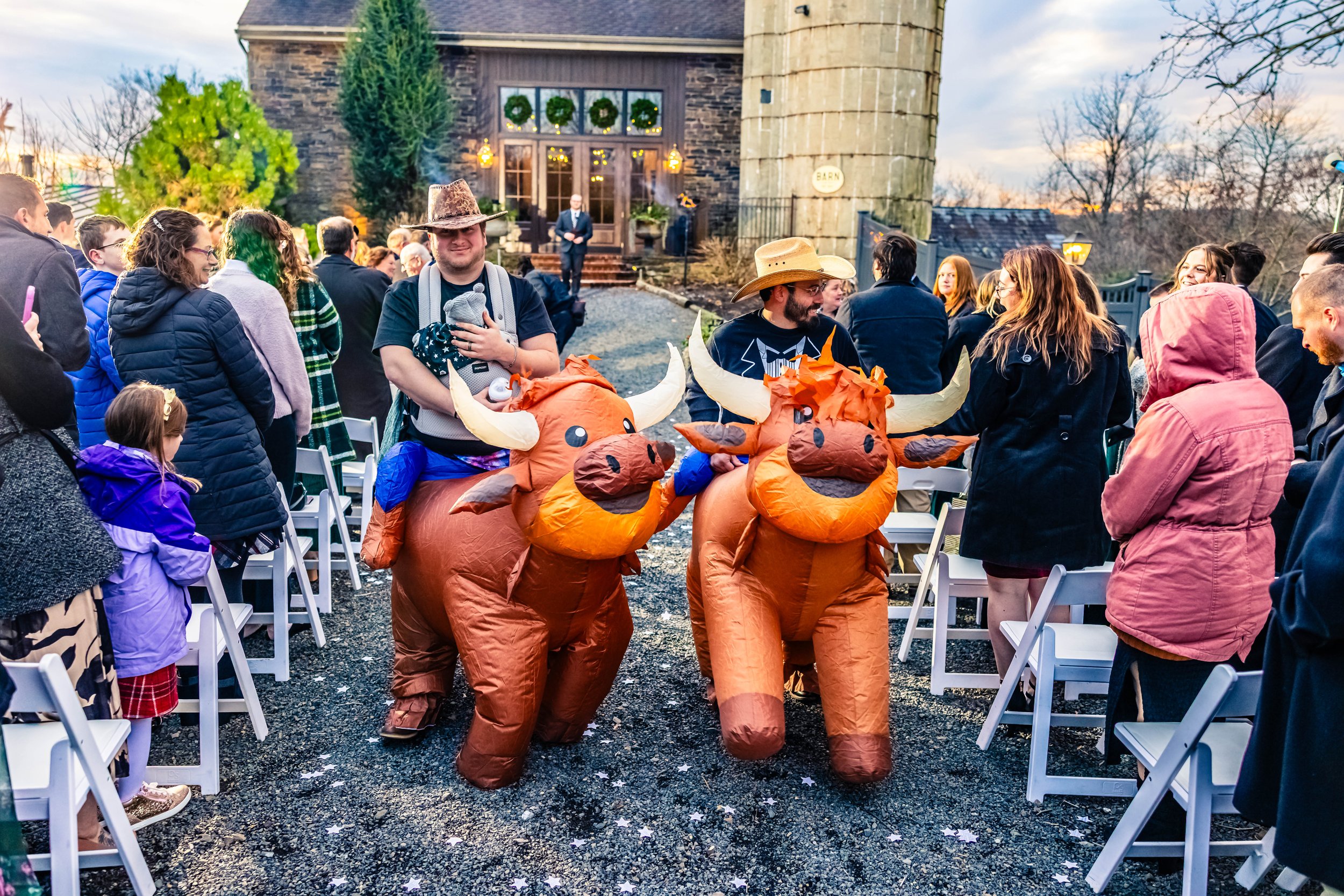 People attending an outdoor event, some wearing cowboy hats, with two individuals in inflatable bull costumes walking among seated guests, a building with festive wreaths in the background.