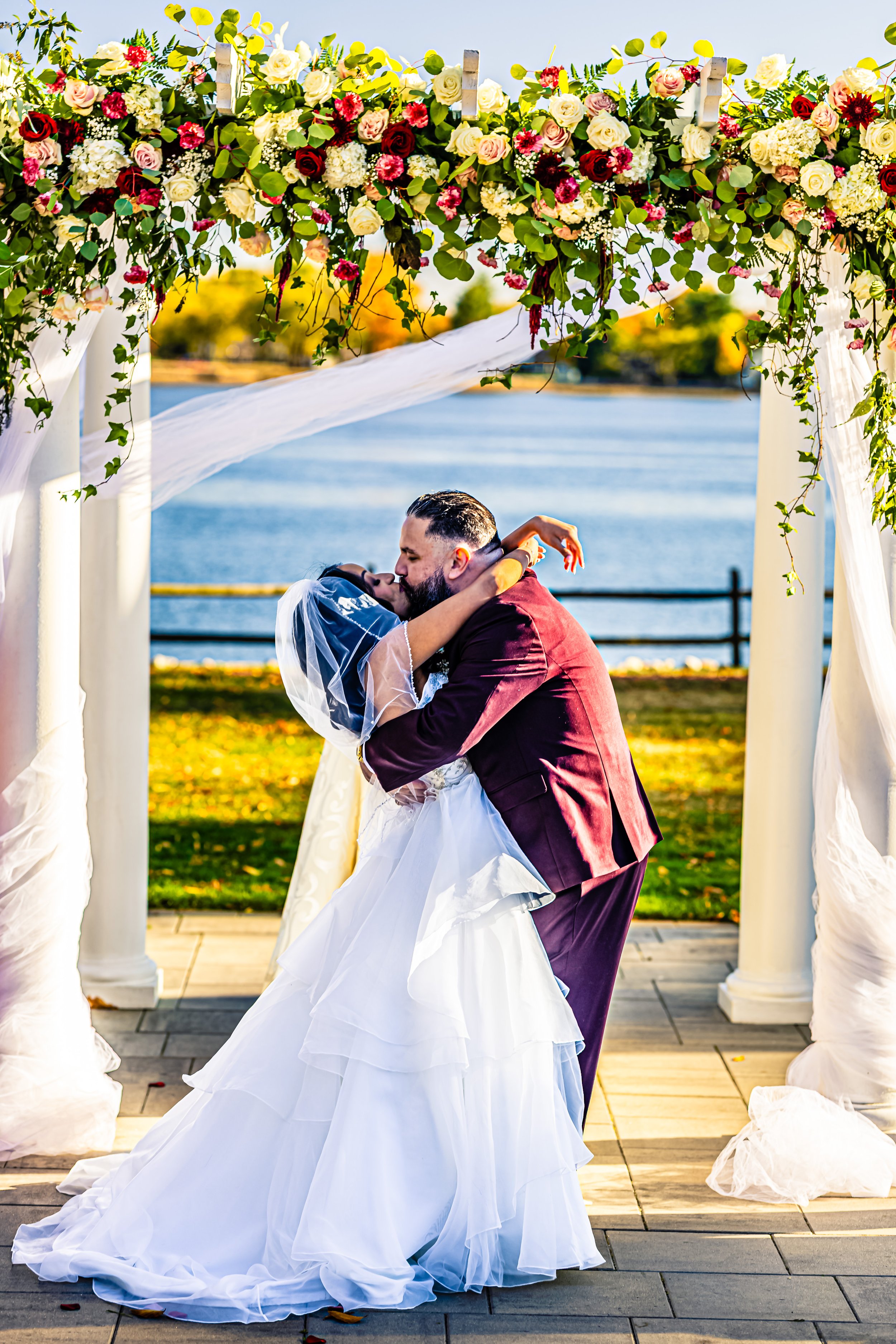 A bride and groom kissing beneath a floral wedding arch by a lake during sunset.