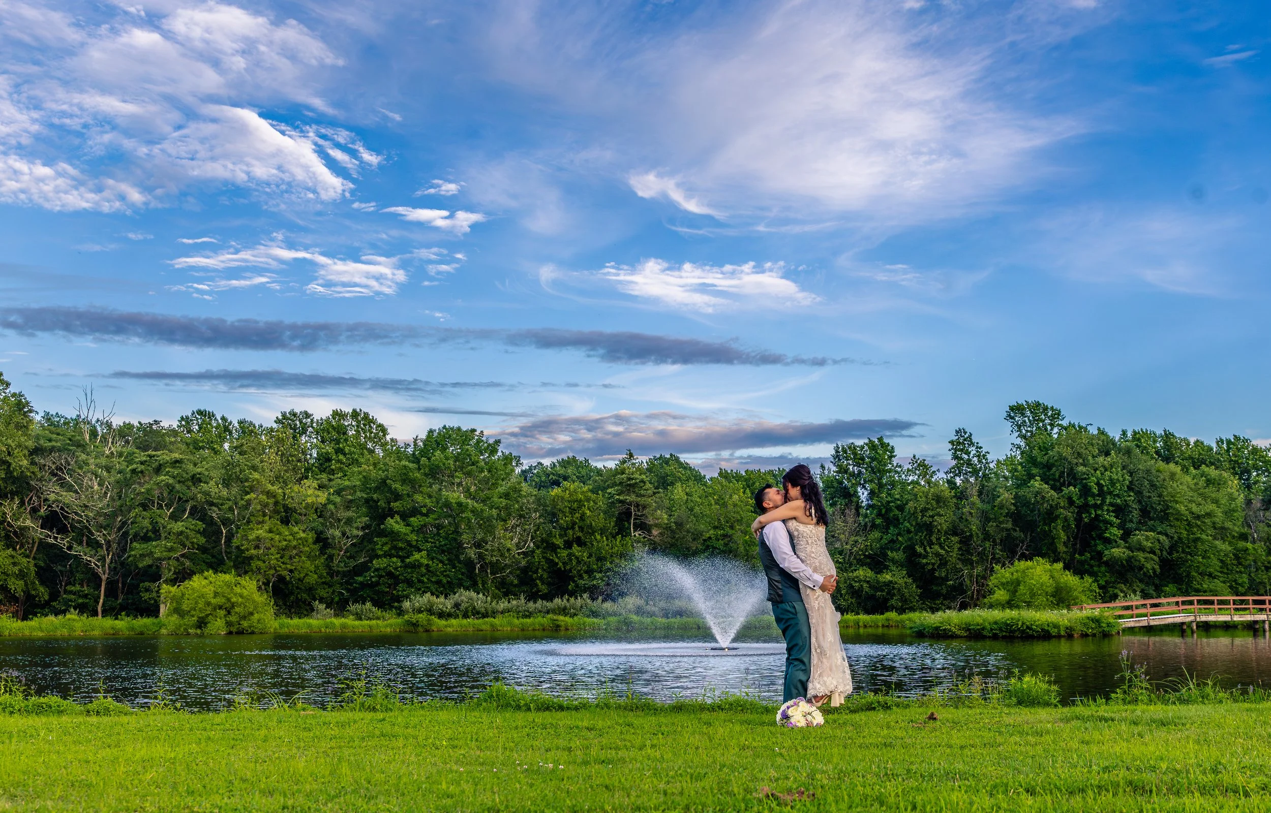 A couple embraces by a pond with a fountain, surrounded by green trees under a blue sky with clouds.