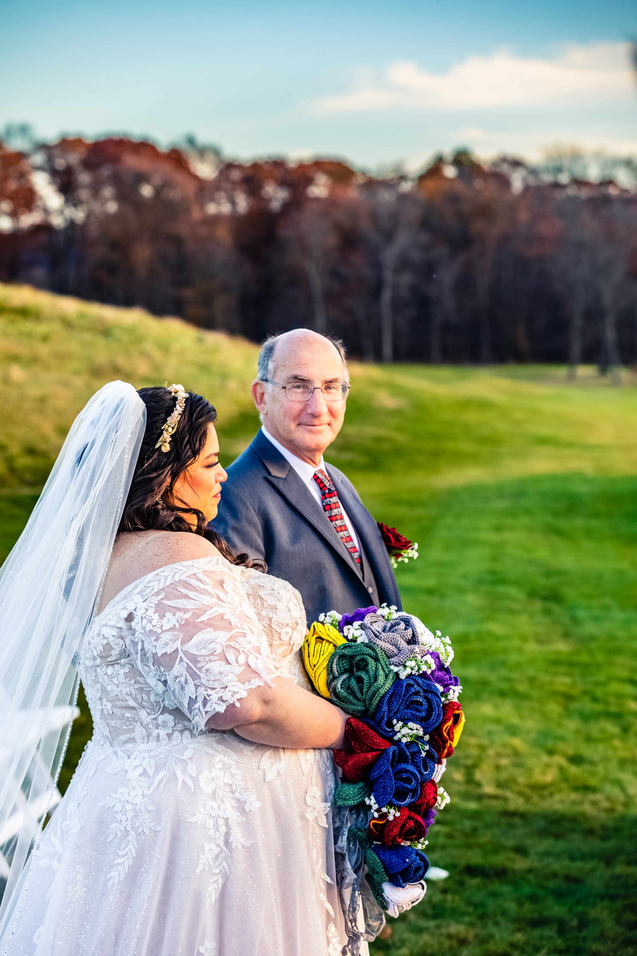 A bride holding a bouquet made of colorful, rolled-up socks standing next to an older man in a suit outdoors on a grassy field with trees in the background.