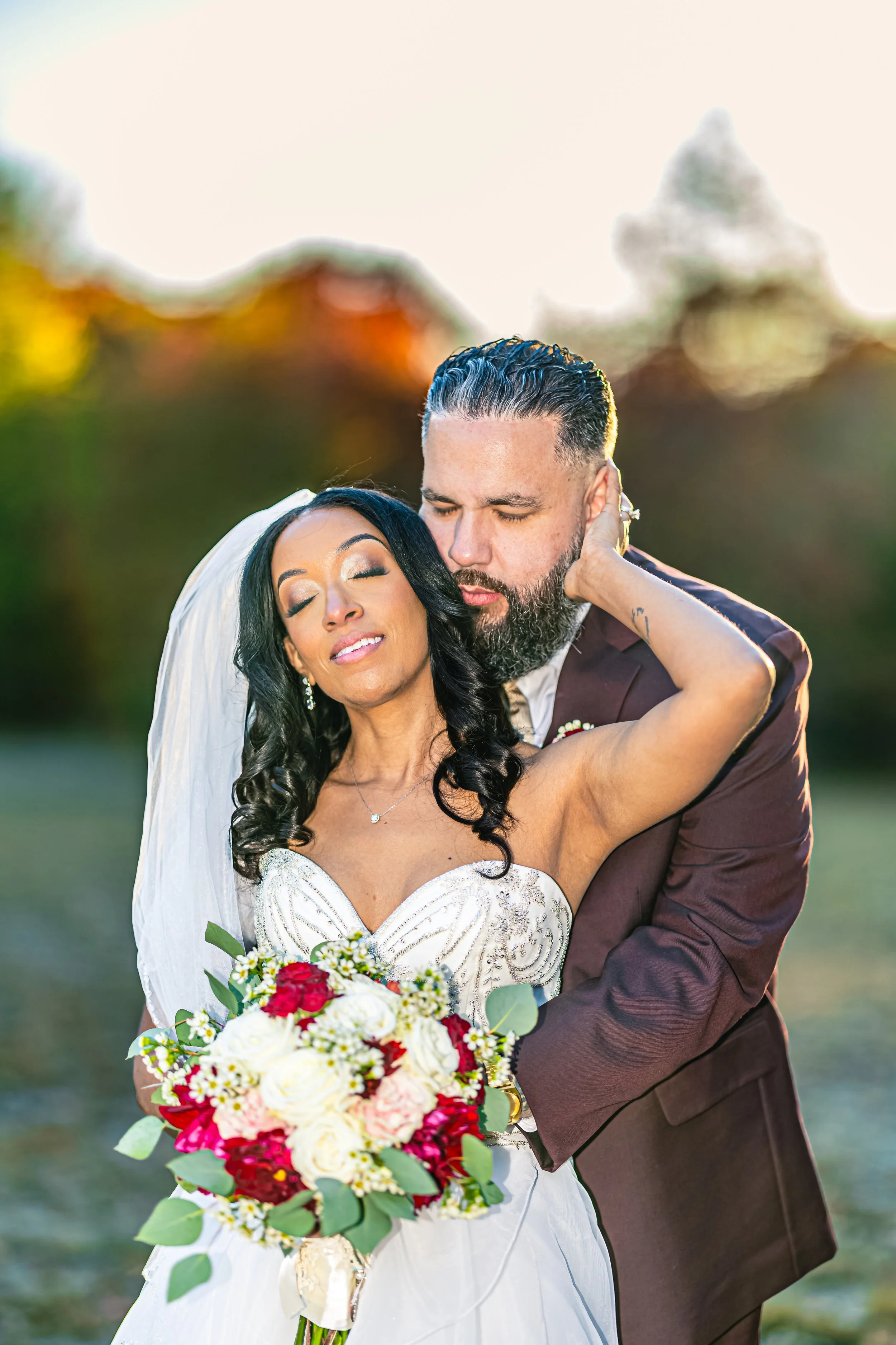 A bride and groom embrace outdoors during sunset, with the bride holding a bouquet of white, red, and pink flowers and the groom gently holding her, both with closed eyes.