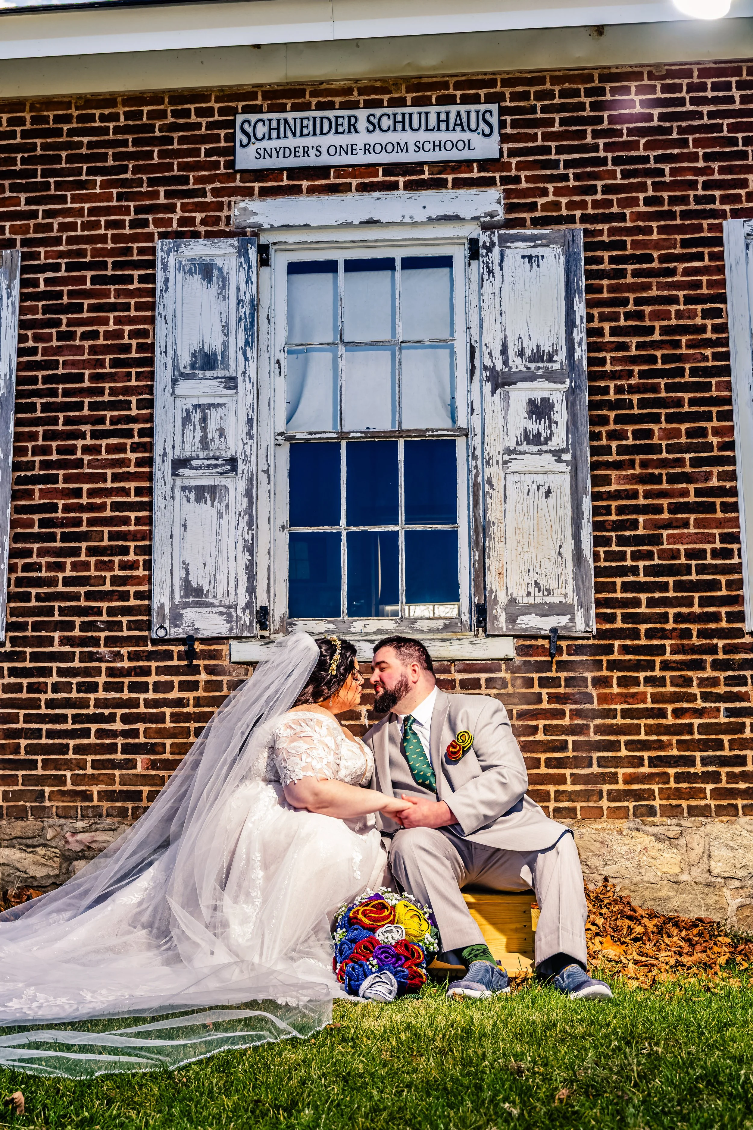 A bride and groom sitting on a wooden crate holding hands and leaning in for a kiss outside a brick school building with an old, weathered window and a sign that reads 'Schneider Schulhaus, Snyder's One-Room School'.