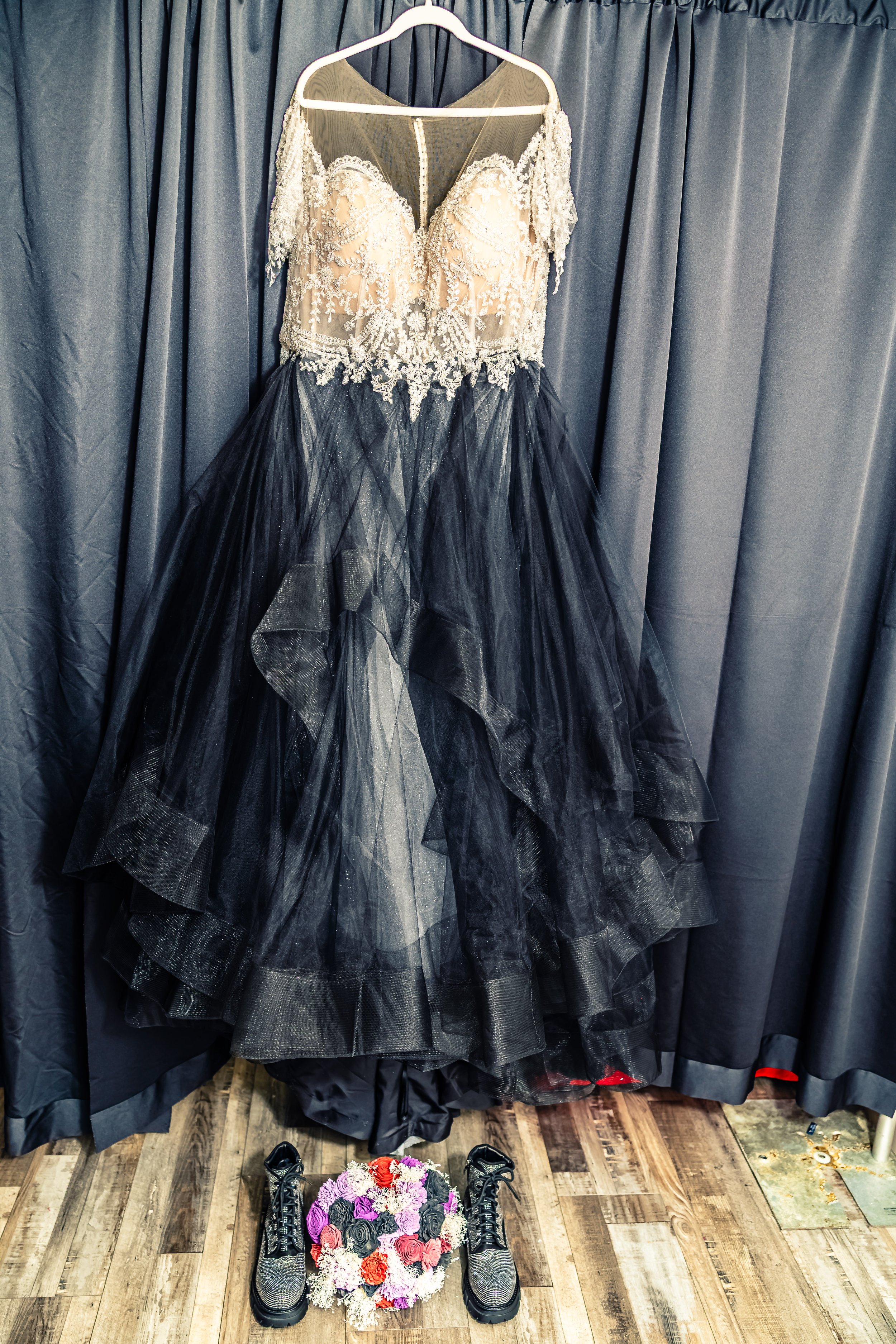 A wedding dress with a lace, beaded bodice and a voluminous black tulle skirt, hanging on a hanger in front of dark curtains. Below, on the wooden floor, there is a bouquet of various colored flowers and a pair of black, glittery shoes.