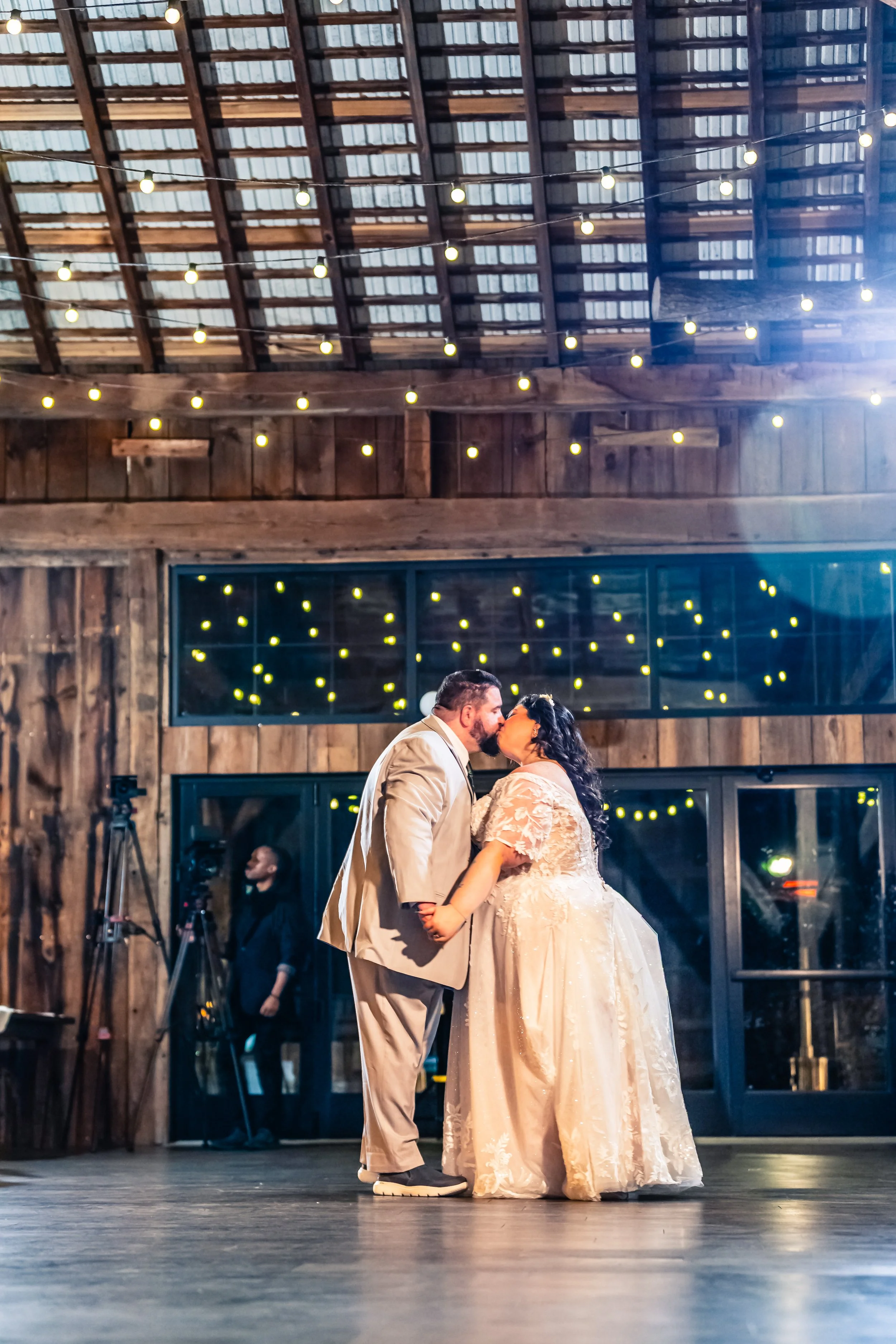 A bride and groom share a kiss during their wedding reception in a rustic barn, with string lights hanging from the ceiling.
