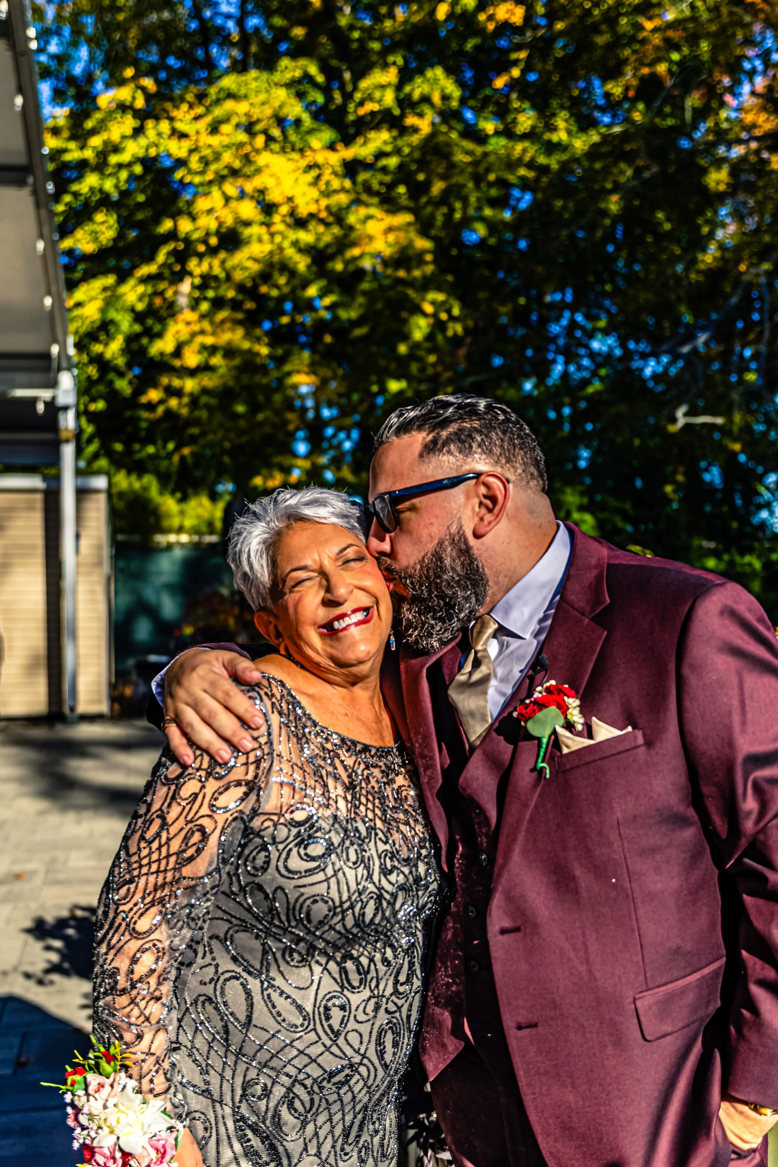 A man in a burgundy suit and sunglasses kisses a woman with gray hair on the cheek during an outdoor event on a sunny day, with trees in the background.