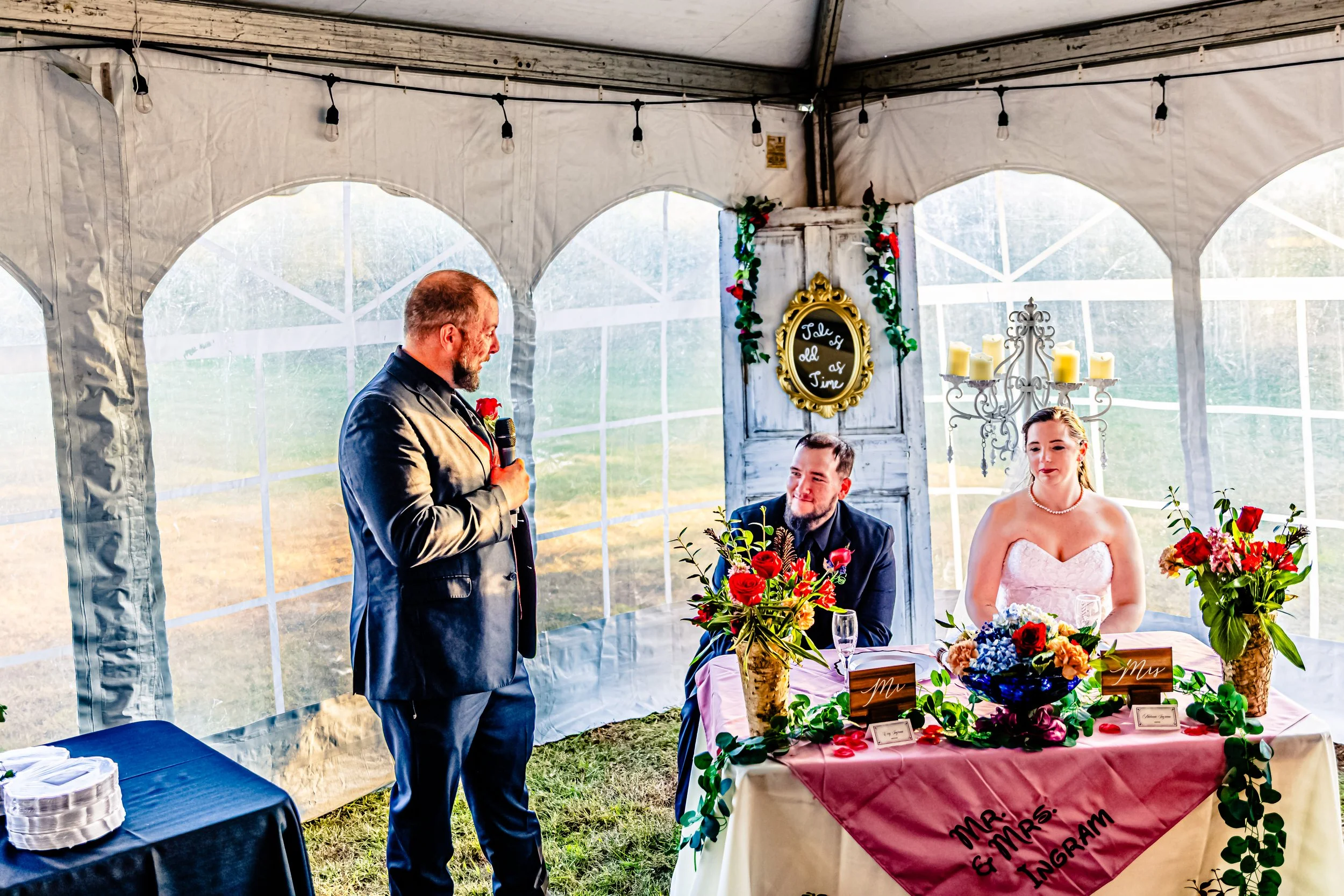 A wedding reception scene inside a white tent with three people: an older man giving a speech with a microphone, a groom seated at a table smiling, and a bride next to him with a serious expression. The table is decorated with flowers and signs readi