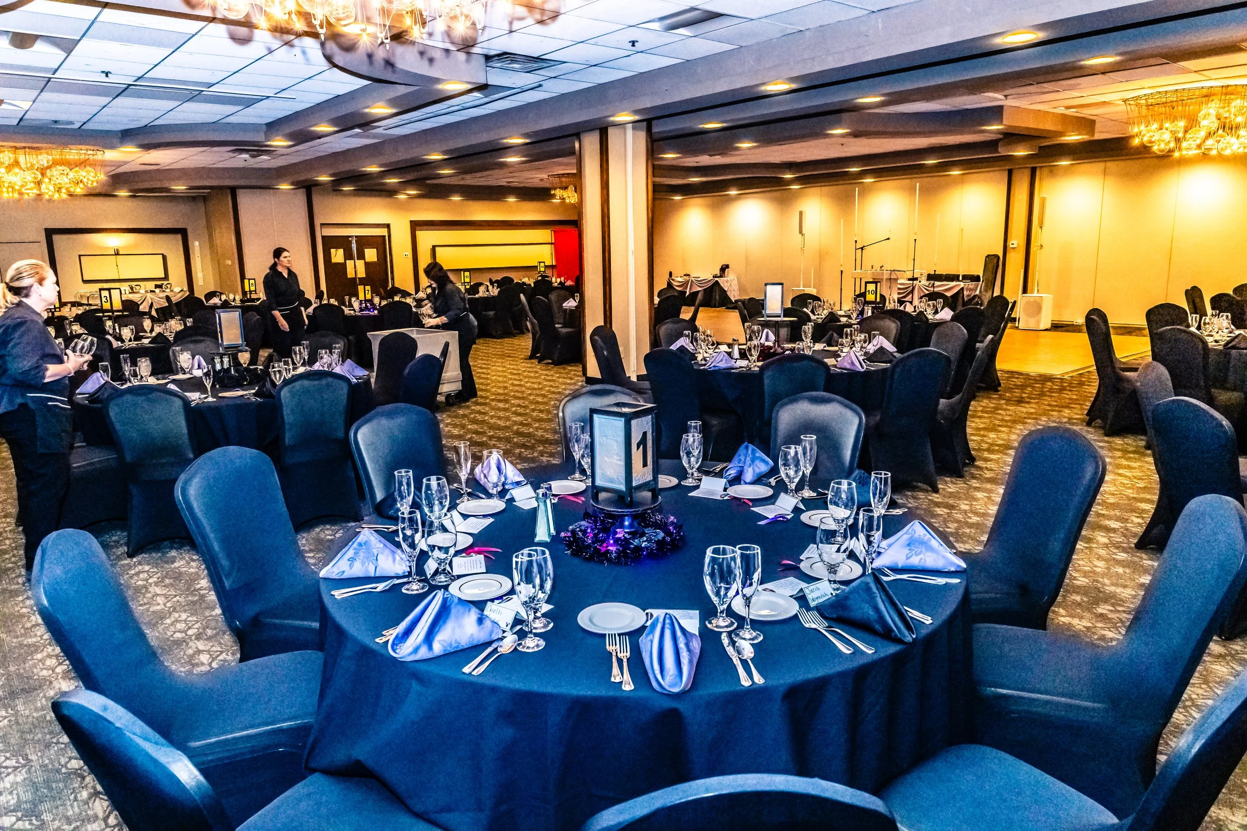Elegant banquet hall with round tables covered in black tablecloths, set with glassware, plates, napkins, and silverware, with stage and musical instruments at the back. Guests and staff are preparing for an event.