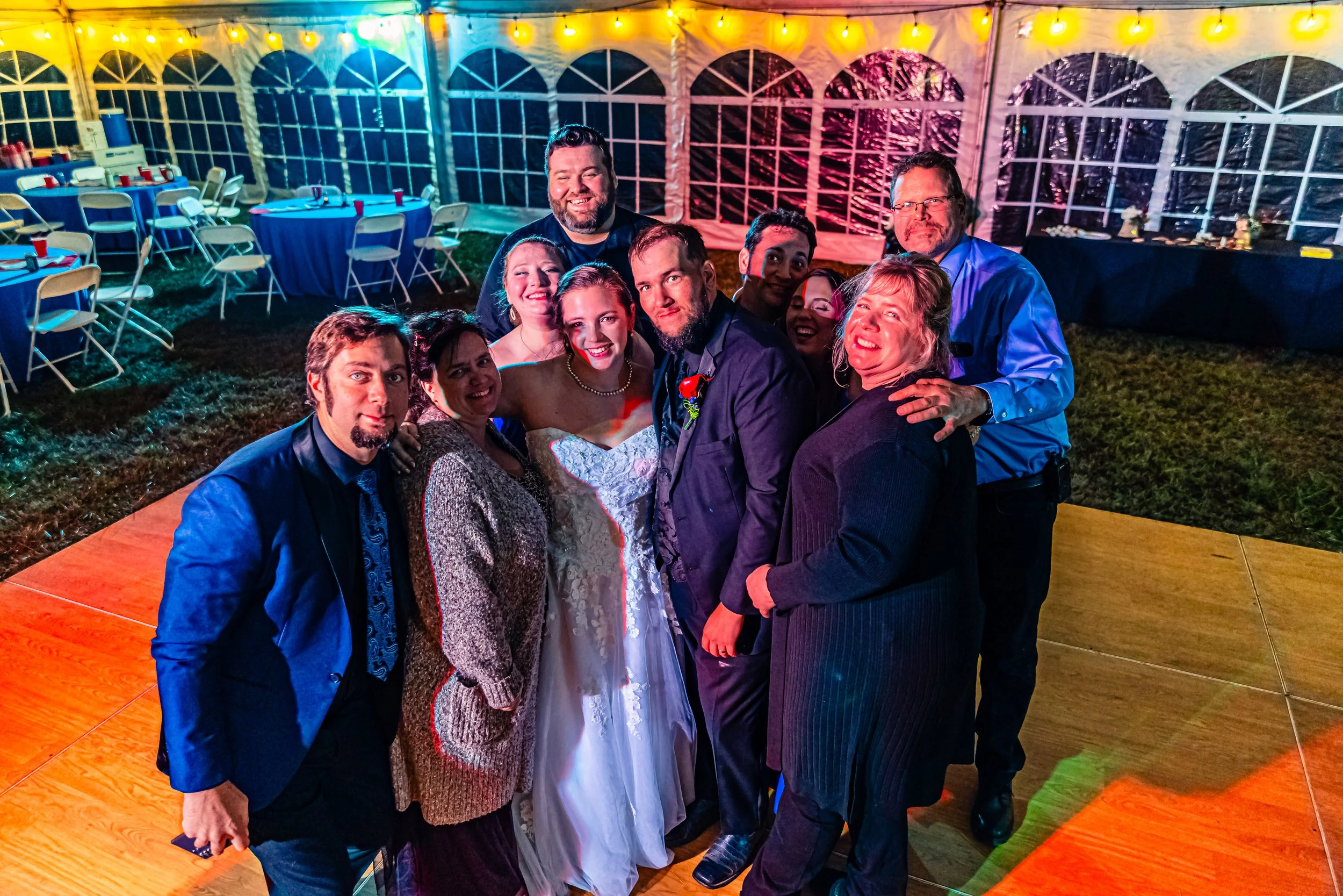 Group of people celebrating at a wedding reception inside a decorated tent with string lights, tables, and chairs in the background.