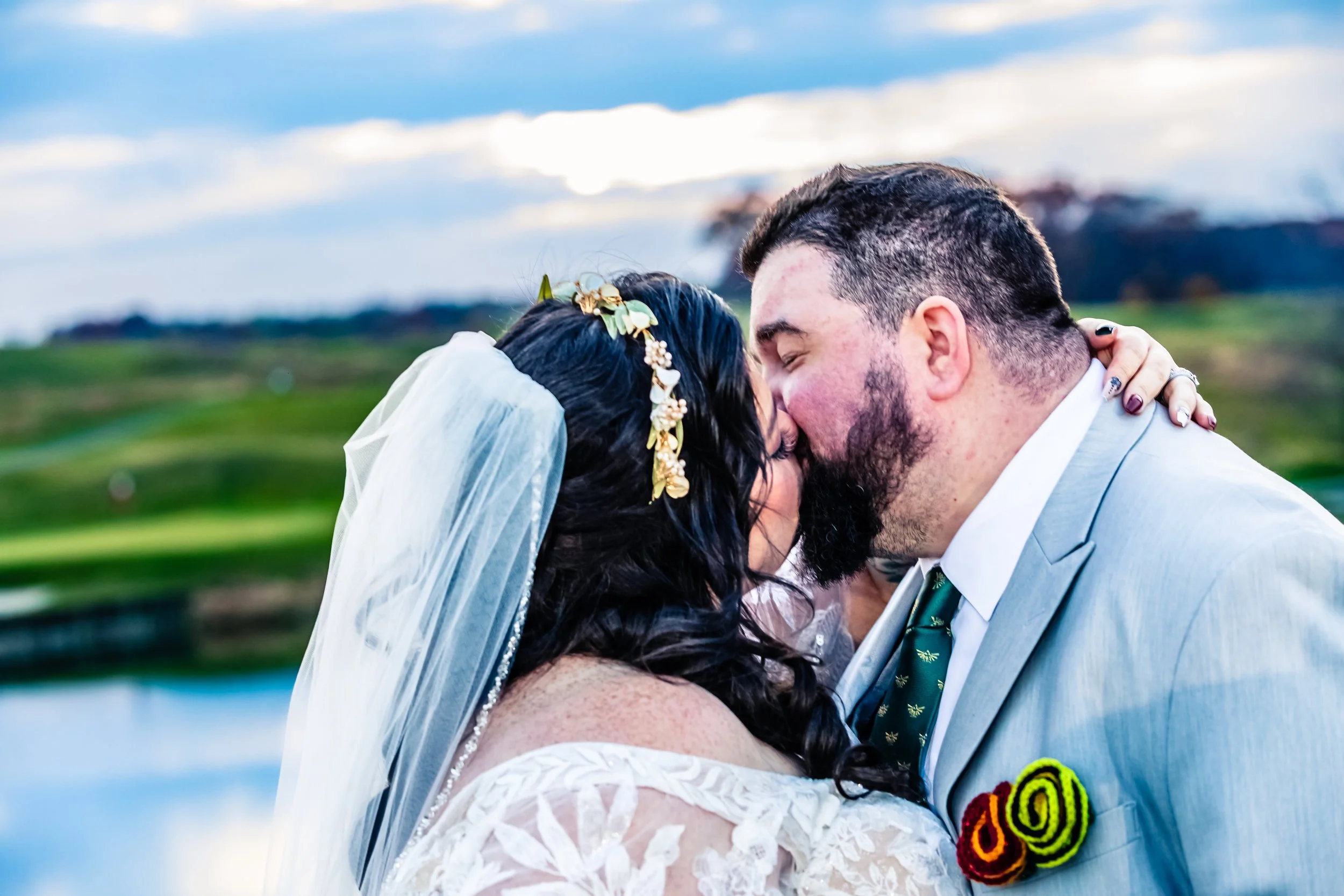 A bride and groom share a kiss outdoors with a scenic landscape in the background, overcast sky, green fields, water, and hills.