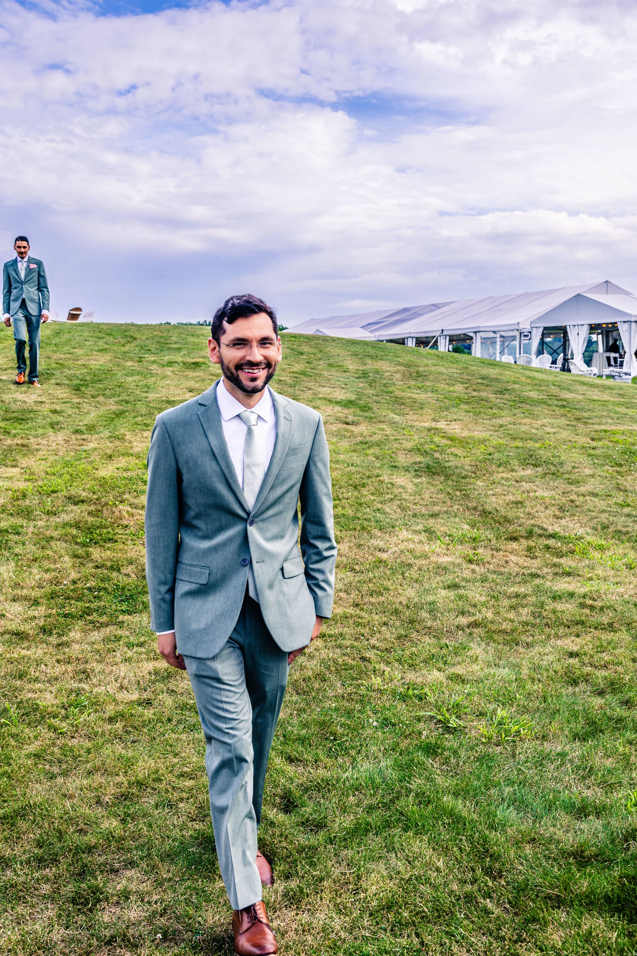 Man in a gray suit walking on a grassy field with another man in a suit in the background near a white tent.