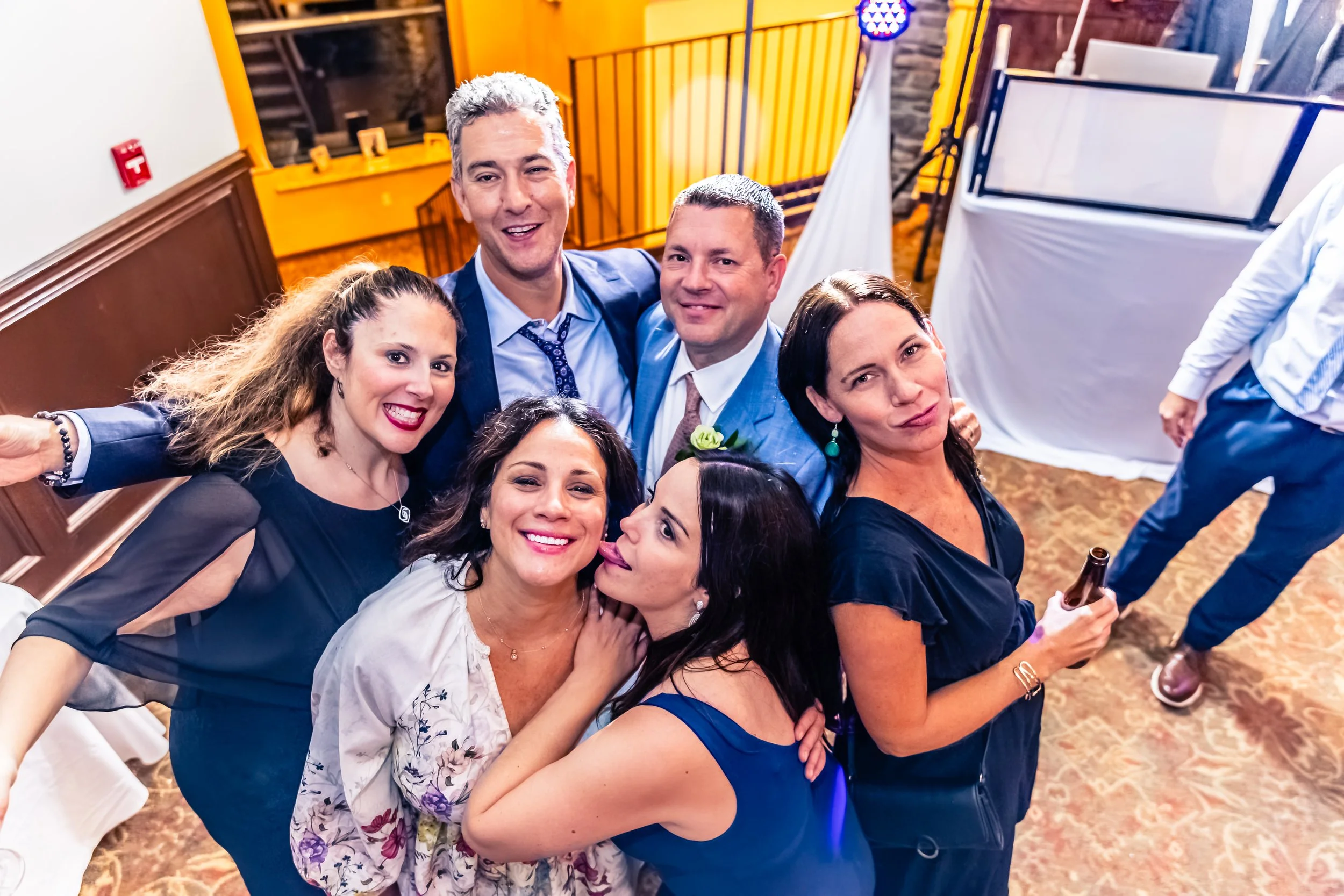 Group of six people at a party, smiling and posing for a photo. Four women and two men dressed in semi-formal attire, standing indoors in a decorated venue with yellow and white drapes or barriers.