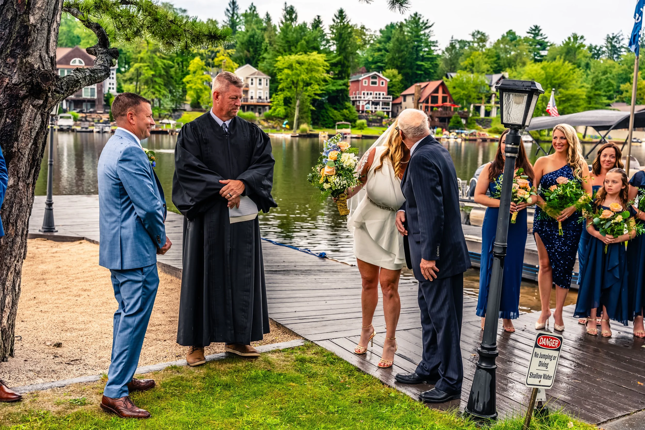Wedding ceremony by a lakeside with officiant, bride, groom, bridesmaids, and groomsmen, with boats and houses in the background.