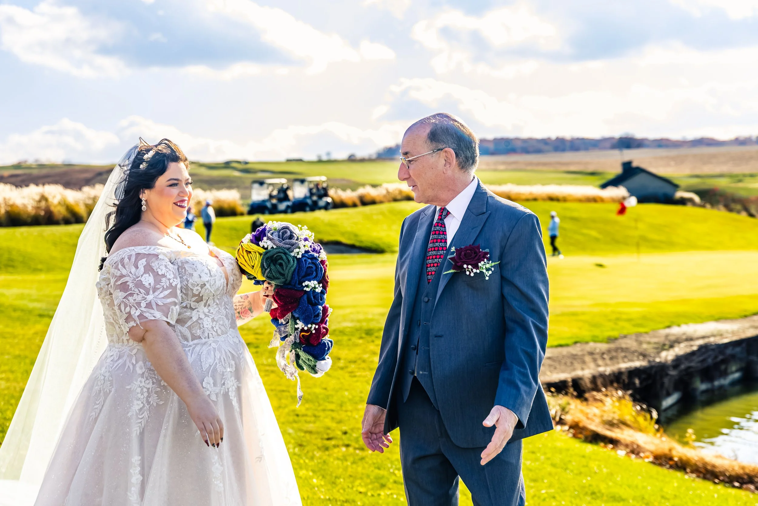 A bride in a white lace wedding dress and veil, holding a colorful bouquet, smiles as she talks to an older man in a gray suit with a boutonniere, on a sunny golf course. There are golf carts and people in the background.