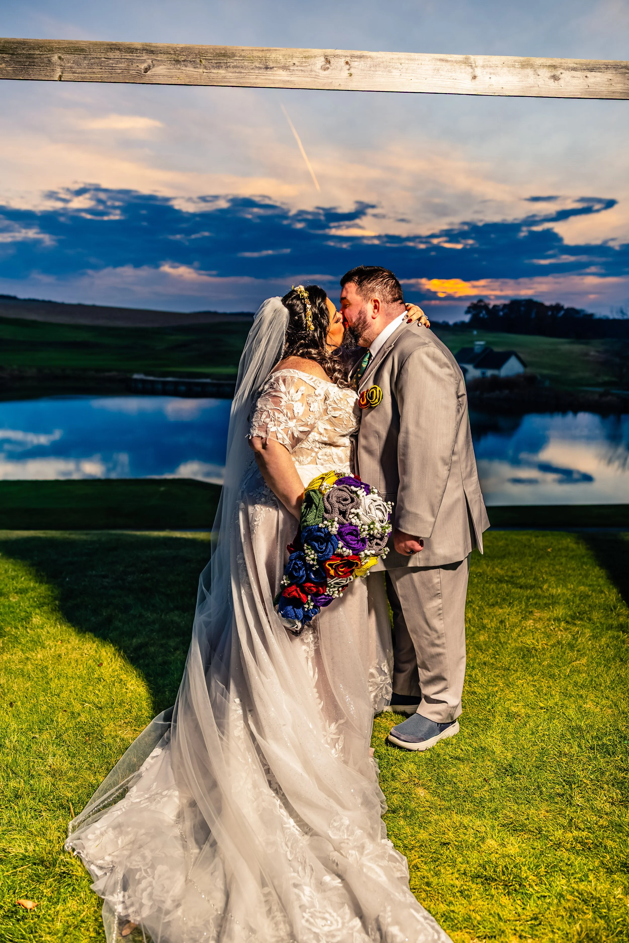 A wedding couple sharing a kiss at sunset near a lake, with a view of the sky and landscape in the background.