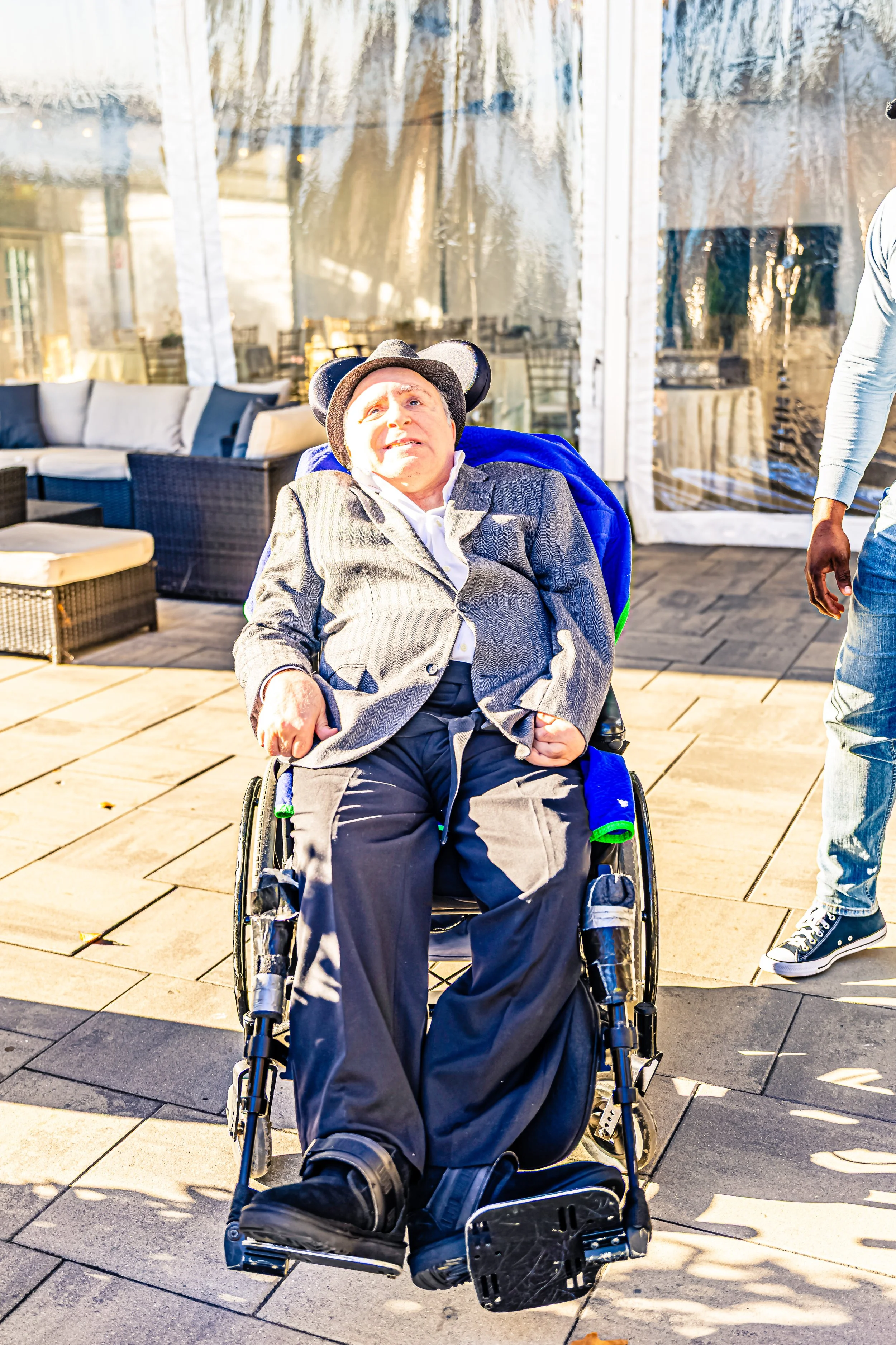 A man in a wheelchair wearing a gray suit and a fedora hat, sitting outside on a patio with wicker furniture and a glass enclosure.
