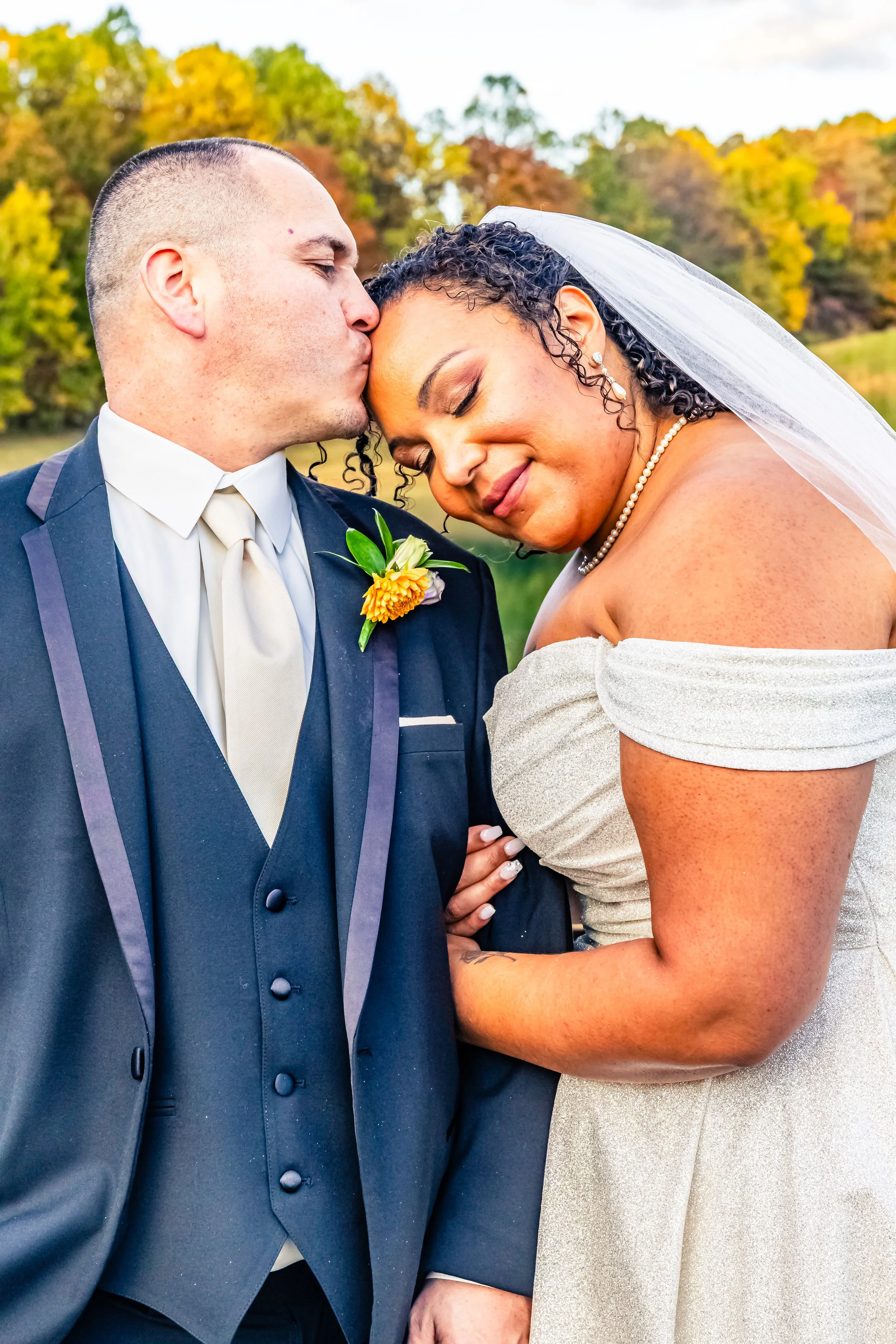 A bride and groom sharing a tender moment outdoors, with the groom kissing the bride on the forehead, surrounded by fall foliage.